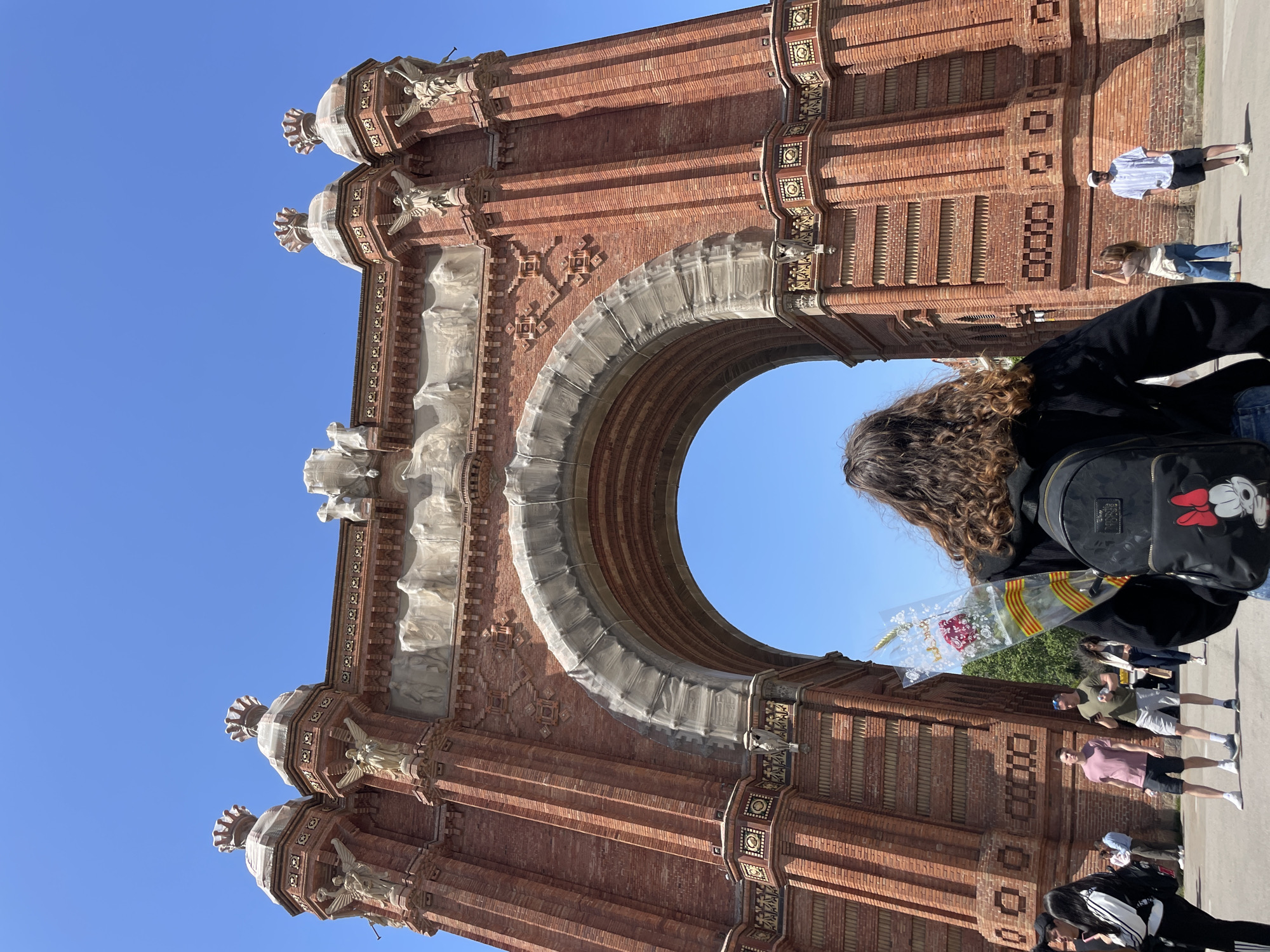 The image captures a sunny outdoor scene in Barcelona, Spain, featuring the prominent Arc de Triomf. The archway is constructed from distinctive red brick in the Neo-Mudéjar style, adorned with intricate details, sculptural elements, and geometric patterns. Parts of the arch, including some sculptures and sections of its inner curve, are covered in white protective material or scaffolding, suggesting ongoing maintenance or preservation work.
In the foreground, with their back to the camera, is a person with long, curly brown hair, wearing a black jacket and a black backpack. The backpack features a white Mickey Mouse head design with red ears and a red bow. This individual holds a clear plastic-wrapped bouquet containing a red flower and a ribbon patterned with yellow and red stripes, indicative of the Catalan flag.
Through and around the archway, several other people are visible, dressed casually, suggesting a public space frequented by visitors or locals. The sky is a clear, bright blue, indicating good weather and strong sunlight, which casts distinct shadows on the brickwork. The overall impression is that of a bustling tourist spot on a pleasant day. No legible text is visible on the archway or surrounding elements, other than the visual branding on the backpack.