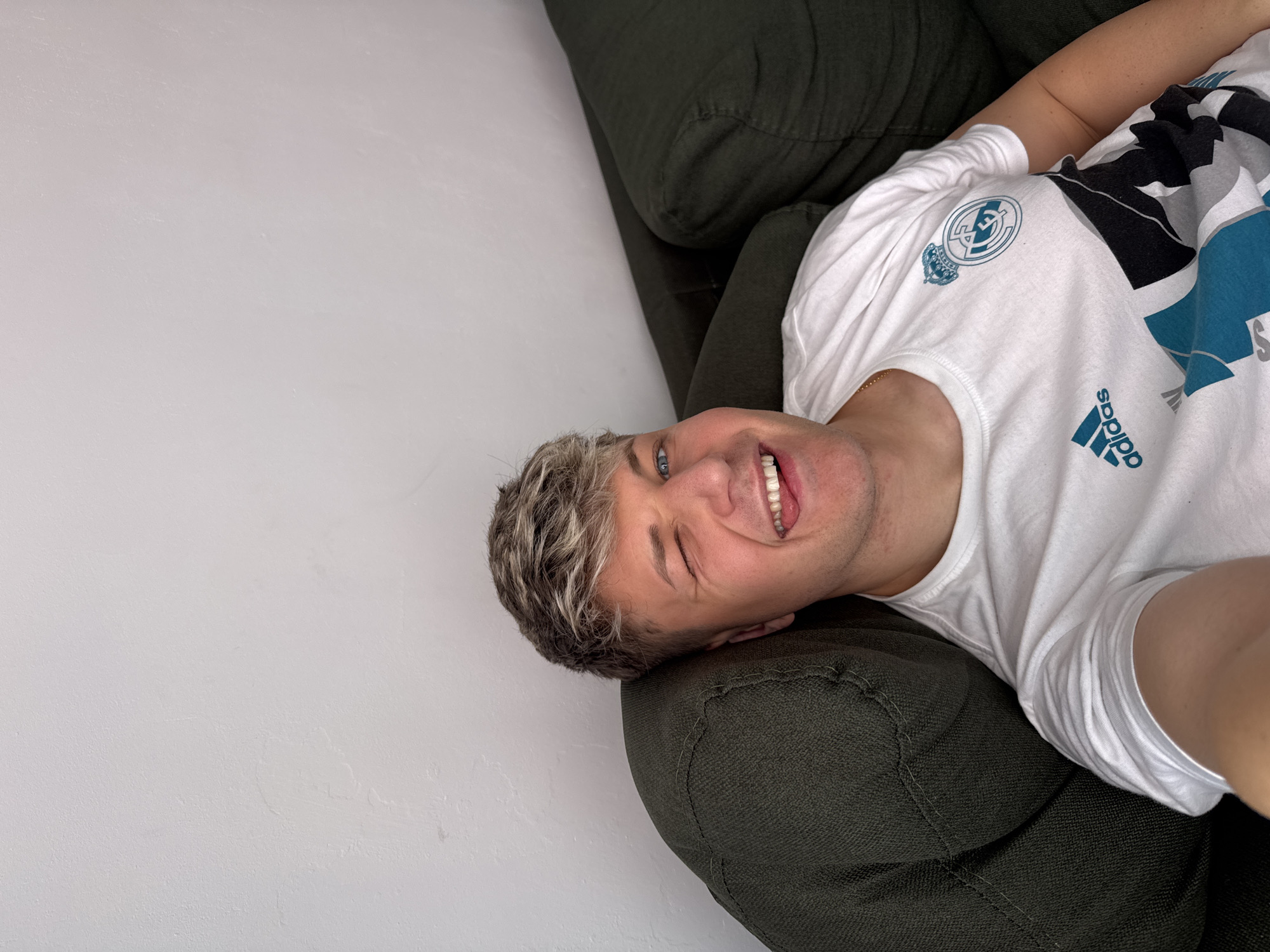 A man with gray and blonde hair, wearing a white t-shirt with the Adidas logo and the Real Madrid logo, is lying on an olive-green couch. He is smiling and winking. The background is a white wall. The location is unknown and cannot be identified as Plovdiv, Bulgaria based on the image.