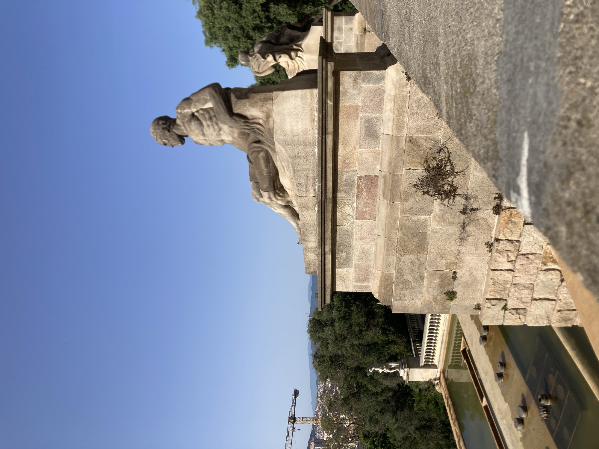 The image depicts a scenic overlook in Barcelona, Spain, likely the Montjuïc hill given the architectural style and the panoramic city view. In the foreground, a large stone statue of a seated male figure, rendered in classical style with detailed musculature, is prominently featured. The statue is positioned on a substantial stone plinth. To the left, a tiered water feature with several small circular fountains is visible, cascading down the slope. Further in the background, lush green trees form a dense canopy, partially obscuring the cityscape. A distant construction crane is also visible on the left side of the image, against the backdrop of a clear blue sky and hazy mountains. Another smaller white statue stands on a pedestal amidst the trees, adding to the classical aesthetic of the location. The overall scene suggests a public garden or a historical monument within the city.