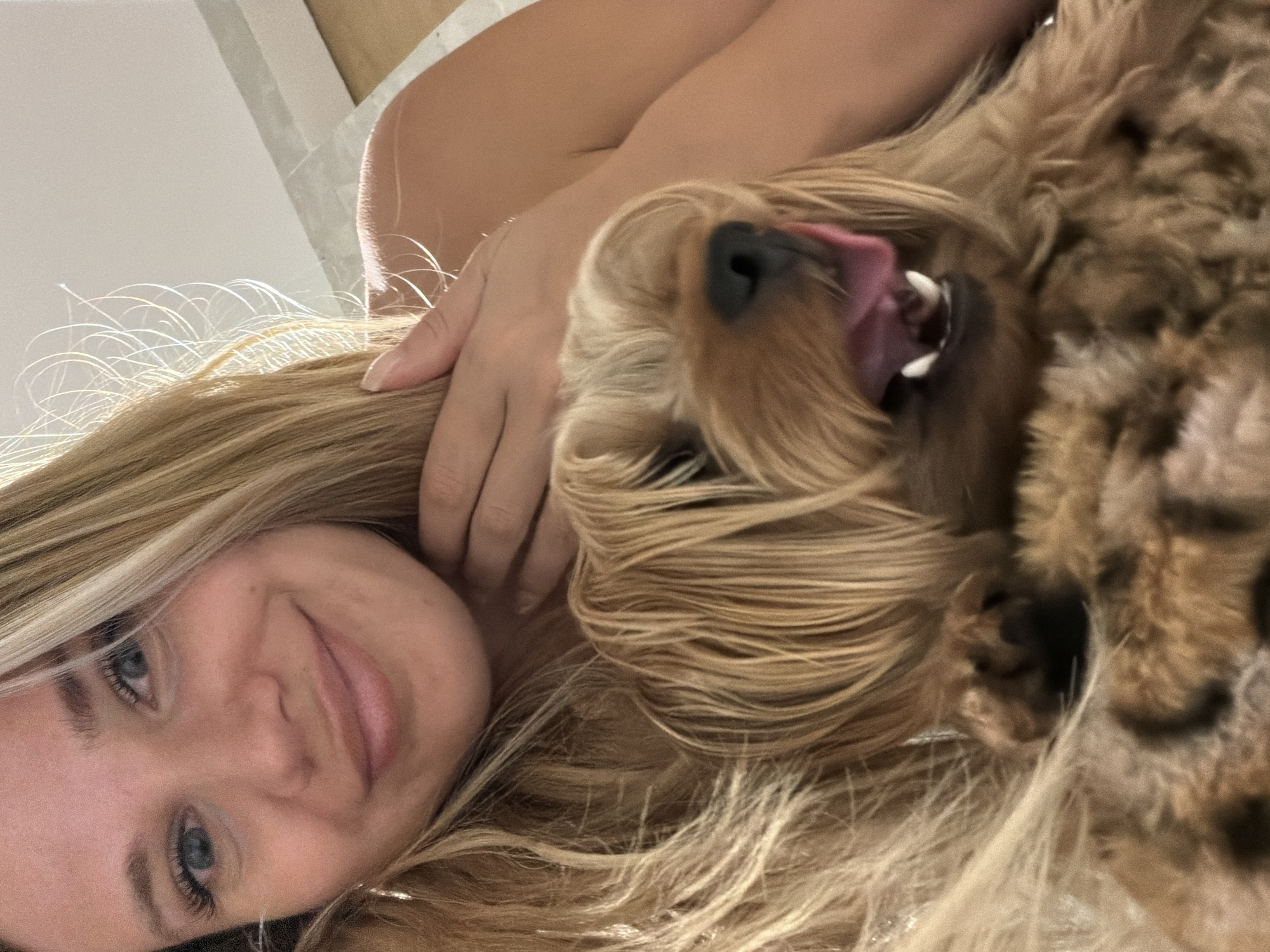 A close-up, low-angle shot features a young woman lying on her side, her head tilted back and looking towards the camera. She has long, blonde hair with lighter streaks that frame her face. Her eyes are blue and appear to be looking up, with visible eyelashes. Her mouth is slightly open, revealing her lips. To her right, a small, tan dog, possibly a Yorkshire Terrier, is also lying on its side, with its head near the woman's. The dog's mouth is open, and its tongue is visible. The background is out of focus, with hints of a white surface and a shadow. The lighting suggests it is daytime, possibly indoors or in a shaded outdoor area. The overall mood appears relaxed and affectionate. There is no visible text in the image.