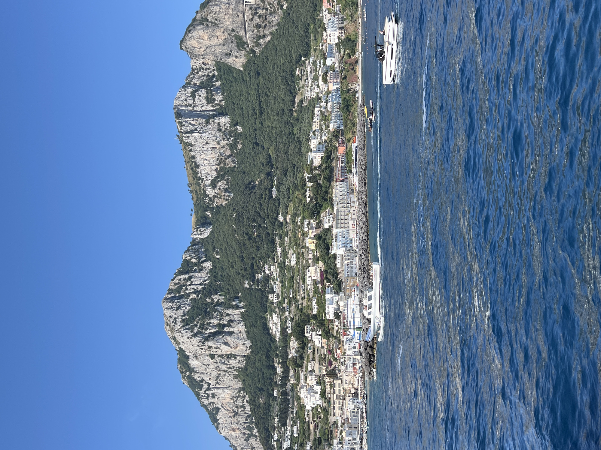 This image captures a scenic view of Capri, Italy, with the distinctive mountainous terrain forming a dramatic backdrop. In the foreground, the deep blue waters of the sea are agitated, with several boats navigating the waves. The most prominent boat is a sleek white vessel in the lower right quadrant, carrying several individuals. To the left, another white boat is moving towards the shore, with more people visible.
The middle ground features the coastline of Capri, characterized by a dense cluster of white buildings with colorful accents, cascading down the hillside. A harbor area is visible, with breakwaters made of dark rocks. The buildings appear to be a mix of residential and commercial structures, with a vibrant urban feel.
The background is dominated by a rugged, grey mountain range, sparsely covered with green vegetation, primarily trees. The peak of the mountain is jagged and imposing, with clear blue sky extending above it. The overall impression is of a sunny day with good visibility, showcasing the natural beauty and developed landscape of the island.