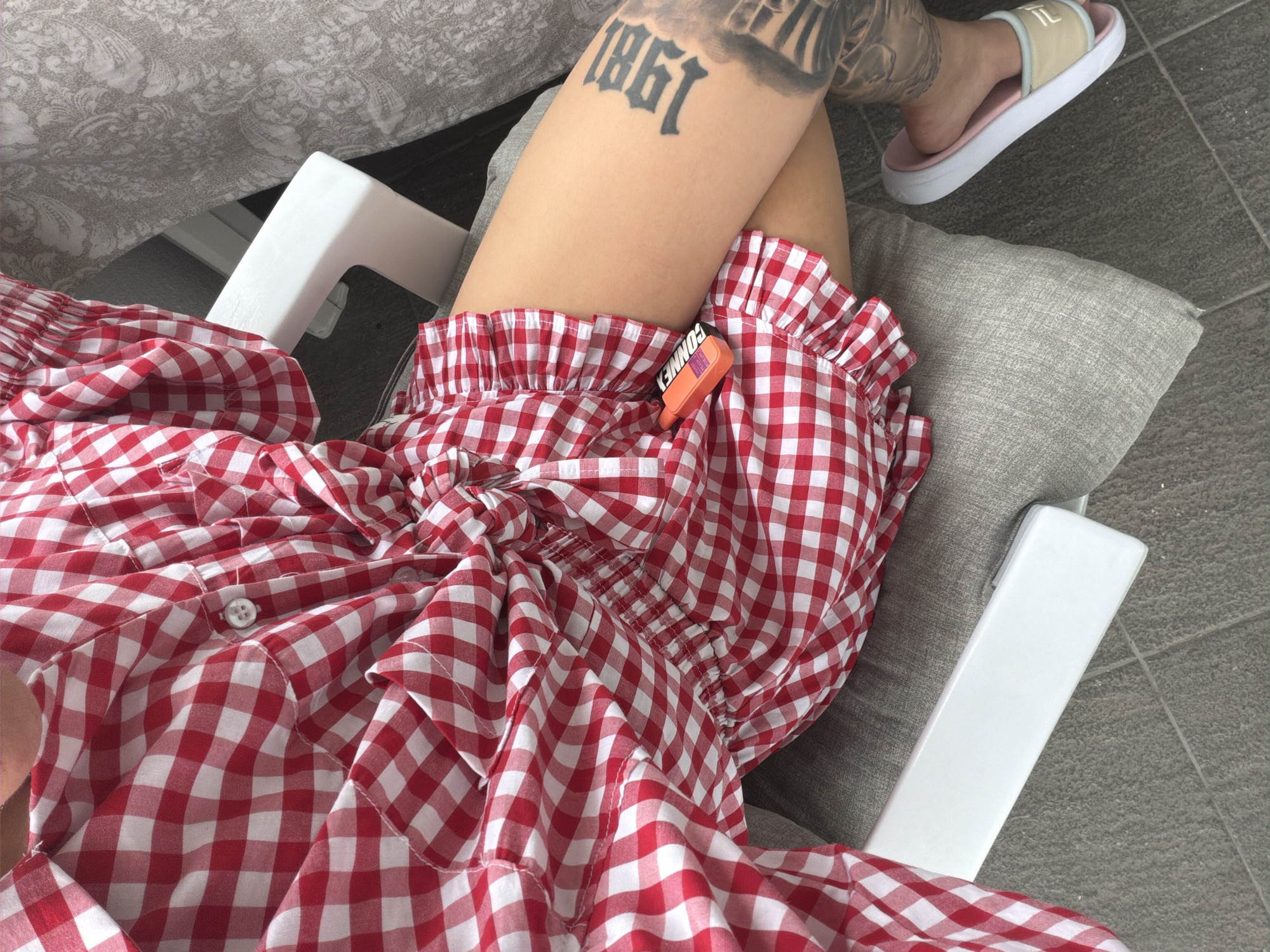 A close-up, high-angle shot captures a person relaxing outdoors, likely on a patio or balcony. The image focuses on the person's lower body, with their legs crossed. They are wearing a red and white gingham-patterned outfit, which appears to be a dress or romper, featuring ruffled edges and a white button closure on the torso.
One bare thigh prominently displays a bold black tattoo of the year "1861", along with other less distinct tattoos further up. On the visible foot, a light beige and pink slide sandal is worn, with the letters "IL" visible on its strap.
Resting between the crossed thighs is a small, bright orange object, possibly a lighter or small gadget, with the word "CONNEY" or "CONNEX" printed in black on its side.
The person is seated on a chair with white armrests. A grey, textured cushion or pillow supports one leg. The background includes a grey patterned fabric, which might be a tablecloth or blanket, and grey tiled flooring, suggesting an outdoor or semi-outdoor setting. The scene conveys a relaxed, warm daytime atmosphere in what could be Plovdiv, Bulgaria.