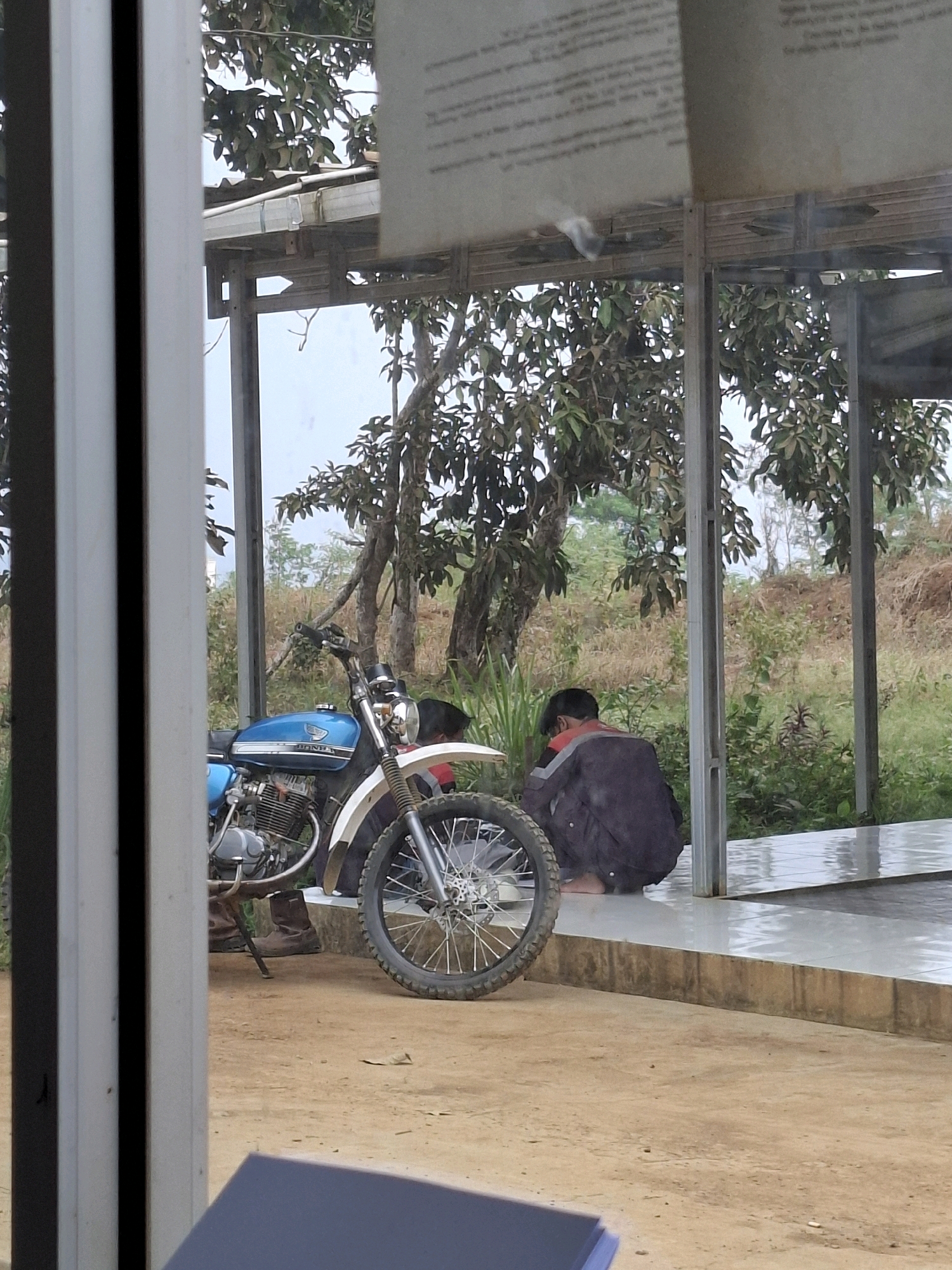 An outdoor scene in Kutasirna, Indonesia, shows a blue and white motorcycle parked on a dirt ground. To the right of the motorcycle, two individuals are seen squatting on a tiled platform. One individual is wearing dark clothing and is positioned facing away from the camera, while the other, partially obscured by the first, appears to be wearing a white garment. The background consists of lush green vegetation and trees under an overcast sky. A portion of a structure with a roof and vertical supports is visible in the foreground.