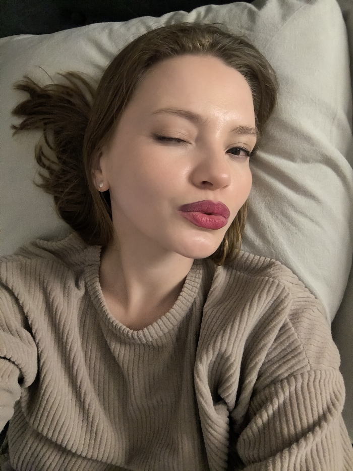 A woman winks, reclining against a pillow. Her light brown hair is tousled, and she wears a beige corduroy sweater. She sports a vibrant red-purple lipstick. A small, round earring is visible on her left ear. The background consists of soft gray pillows. The lighting appears dim.