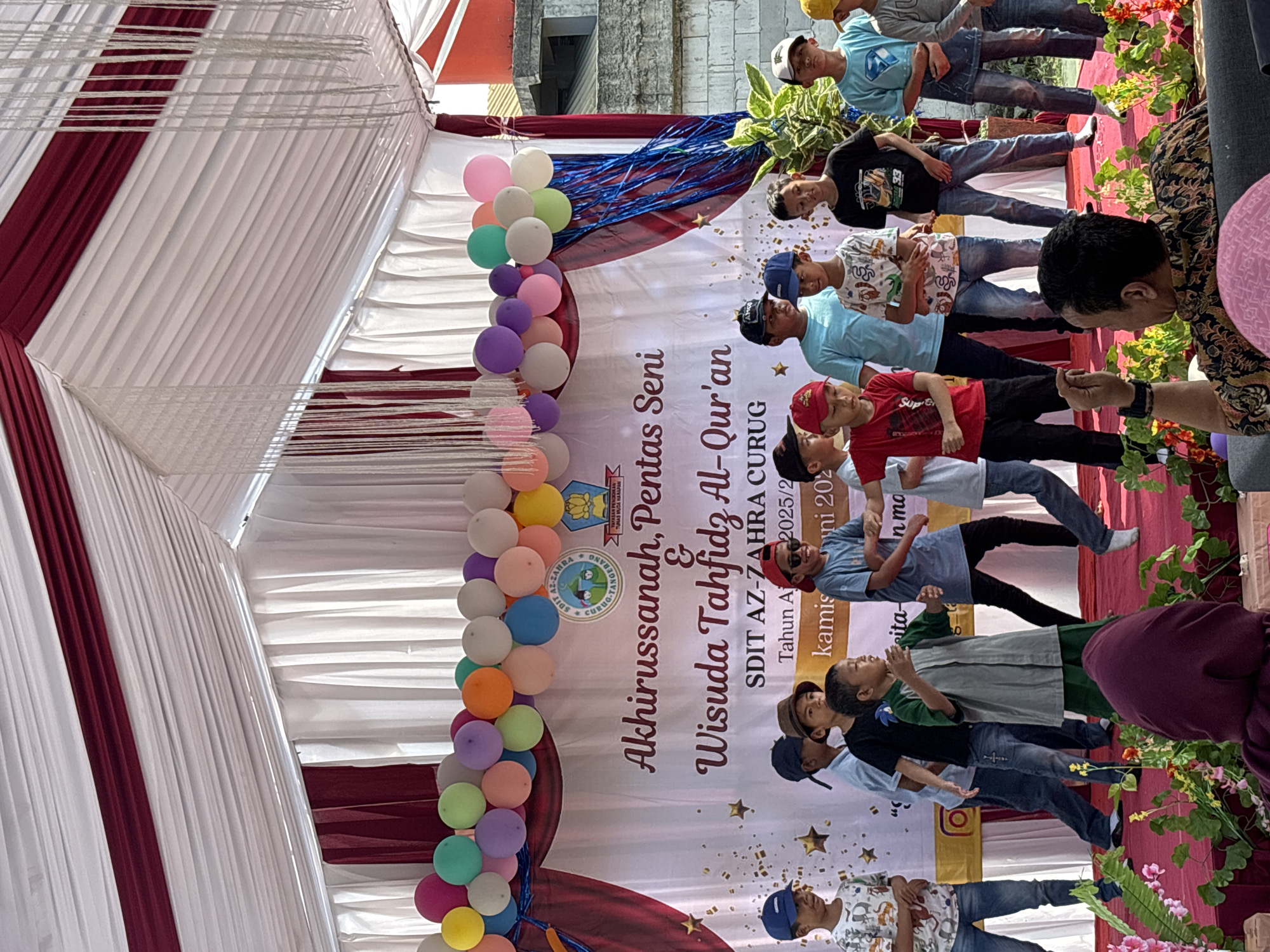 The image depicts an outdoor event, likely a school performance or graduation ceremony, held under a white and maroon tent with hanging decorative ropes. A banner in the background displays text in Indonesian, including "Akhirussanah, Pentas Seni & Wisuda Tahfidz Al-Qur'an" and "SDIT AZ-ZAHRA CURUG". A group of young boys, dressed in casual attire and some wearing caps, are standing on a stage decorated with red carpet and floral arrangements. Some of the boys are actively performing, with one in the center gesturing with his hands and another to his right appearing to sing or speak. The audience is partially visible in the foreground, with one person in a patterned shirt turned away from the camera. Balloons are strung along the top of the stage area. The overall impression is of a vibrant and celebratory occasion taking place in a public or school setting.