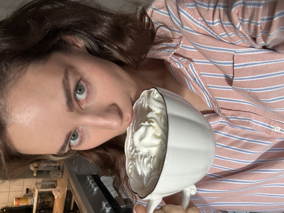 A female subject, approximately 20-35 years old, with brown wavy hair and green eyes, occupies the foreground, looking directly into the camera. She holds a white, fluted ceramic mug with a black rim, filled to the brim with white whipped cream, positioning her mouth to consume its contents. Her attire consists of a pink and light blue vertically striped button-up shirt. The background reveals a domestic kitchen setting, slightly out of focus. Visible elements include a stainless steel appliance (likely a stove or oven) with control knobs, a light-colored countertop, and a shelf unit holding miscellaneous items such as a yellow-and-red container, glass jars, and a speckled mug. The location is an interior residential kitchen within the city of Unknown, Unknown.