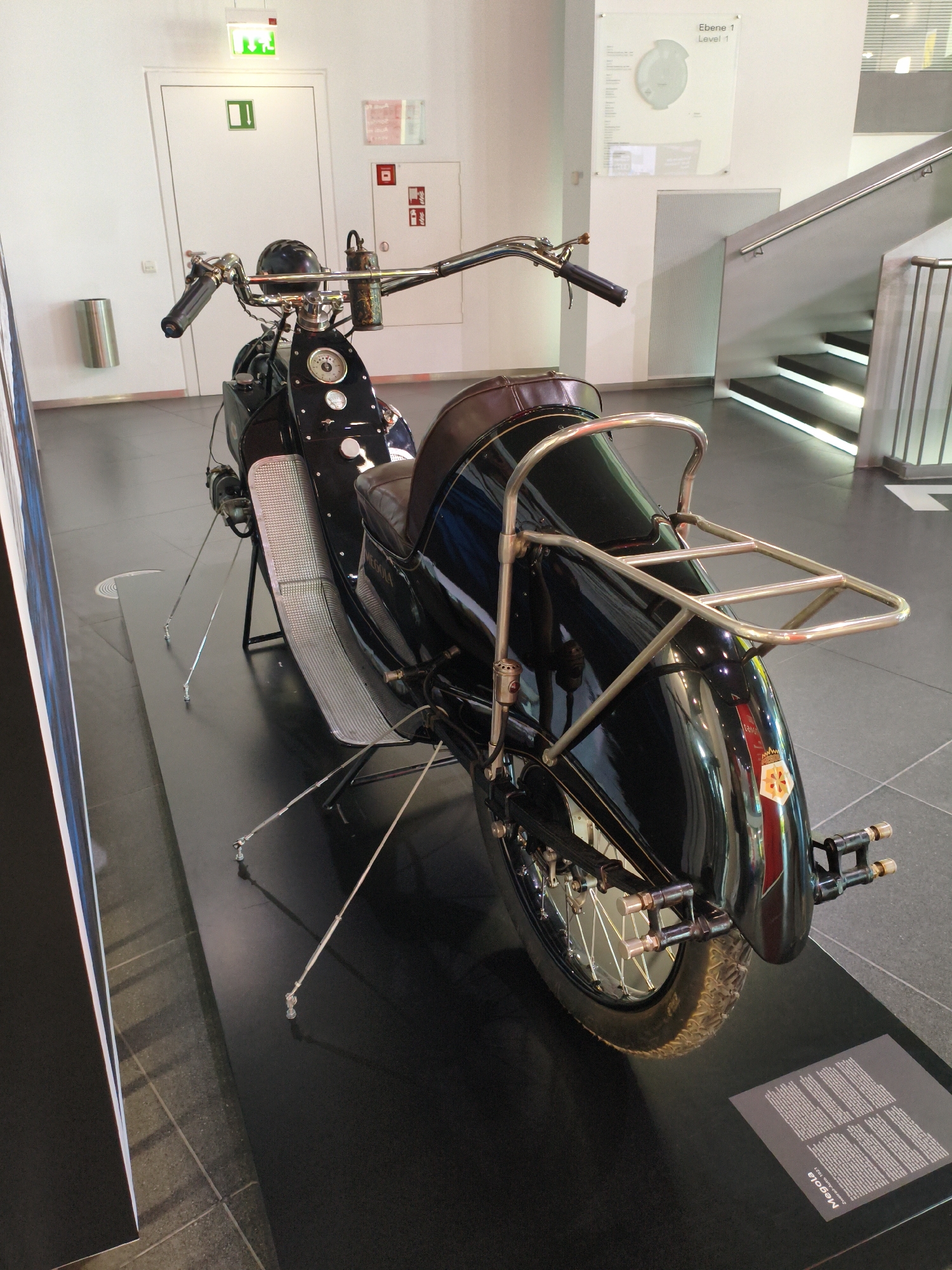 The image shows a vintage black motorcycle with a brown leather seat and chrome accents. The motorcycle is displayed on a black platform in what appears to be a museum or exhibition space. In the background, there is a white wall with a green illuminated exit sign and a white door. To the right, a modern staircase with illuminated steps leads upwards. A glass panel with information about "Ebene 1 Level 1" is mounted on the wall above the staircase. The motorcycle has a luggage rack on the rear. A small descriptive plaque is visible on the platform in the foreground, reading "Megola Zweiradkrad, 1924". This motorcycle is part of the permanent exhibition at the Audi Forum in Ingolstadt, Germany, specifically within the Museum Mobile, which showcases the history of Audi and its predecessor companies.