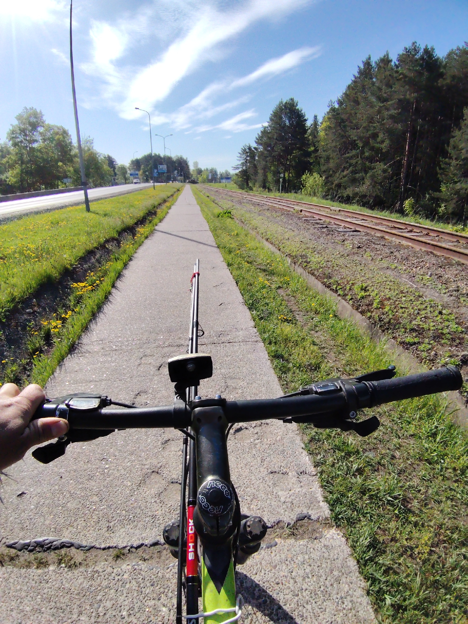 The image captures a first-person perspective from a bicycle rider on a concrete sidewalk in Chaikovsky, Russia. A fishing rod is mounted on the bicycle. The path is flanked by a road with grassy vegetation on the left, and a set of train tracks with denser vegetation to the right. The sky is partly cloudy with sunlight. The bicycle's handlebars and part of the frame are visible. The rider's hand is on the left handlebar.