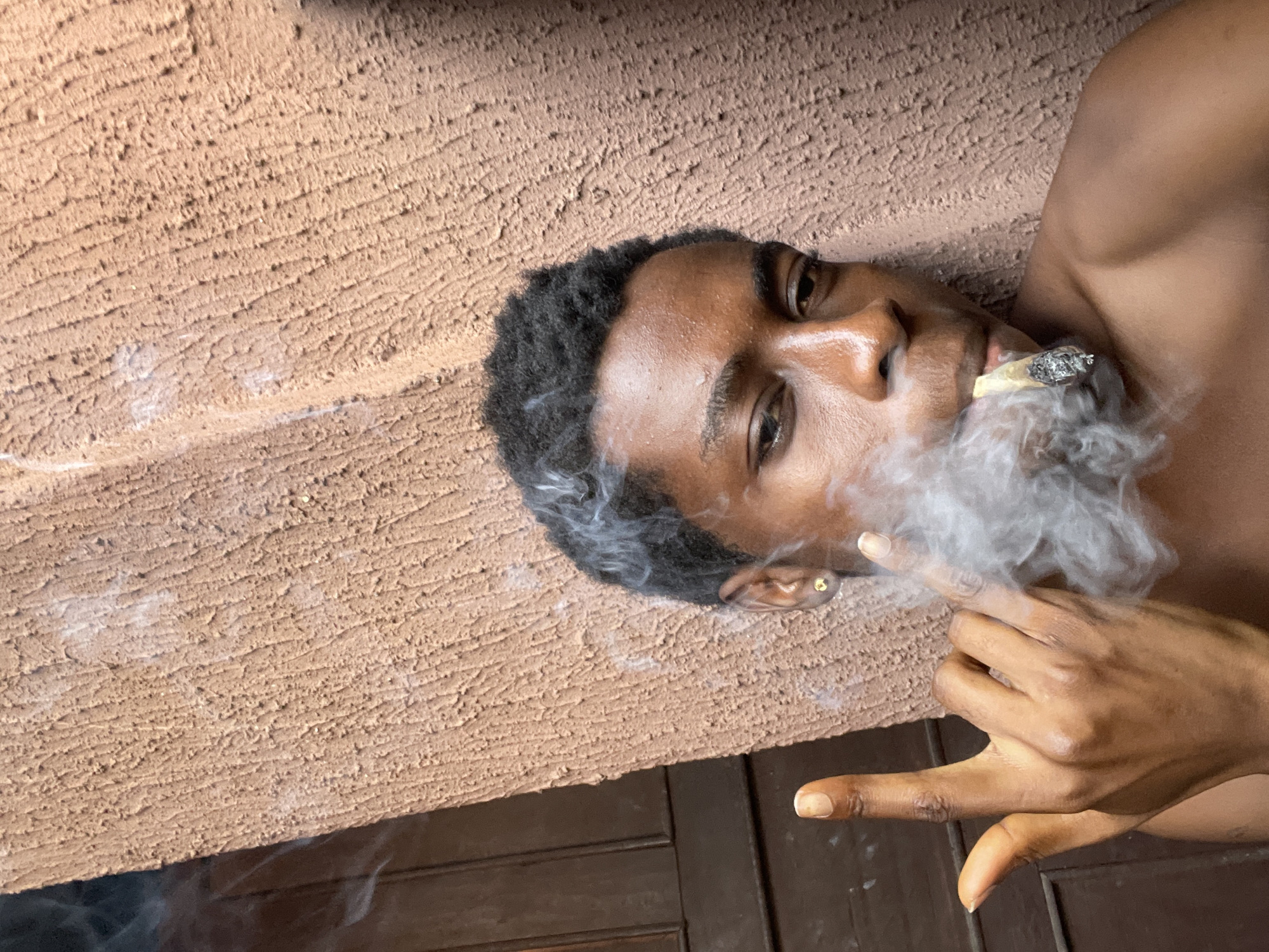 A dark-skinned person lies on their side, angled upwards in the frame, exhaling smoke from a lit object held between their lips. The person has short dark hair and a small gold earring. Their right hand is visible, making a hand gesture. The background features a rough, tan-colored wall and dark wood. The location is identified as Nkpor, Nigeria.
