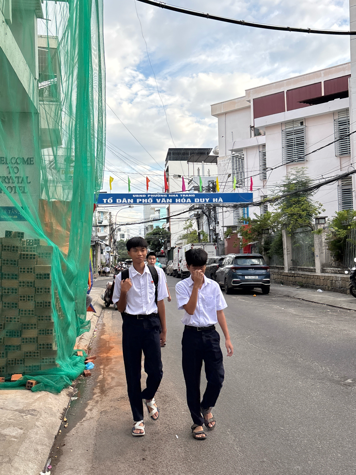 Two young males wearing white shirts and dark pants walk down a street in Nha Trang, Vietnam. One male is in the foreground, facing mostly forward, with his left hand near his face. The other male walks slightly behind and to his right. They are both wearing sandals and carrying backpacks.
The street is paved with asphalt and lined with buildings. On the left side of the street, there is a construction site covered with green netting, with stacks of bricks visible. A sign on the left reads "WELCOME TO CRYSTAL HOTEL". Above the street, there is a blue banner with Vietnamese text which translates to "UBND PHUONG HOA TRANG TO DAN PHO HOA DUY HA". Colorful flags are strung across the street.
Vehicles, including a dark SUV with license plate "79A-641.89", a white truck, and motorcycles, are parked or driving on the street. Trees and electrical wires are also present. The sky is partly cloudy.