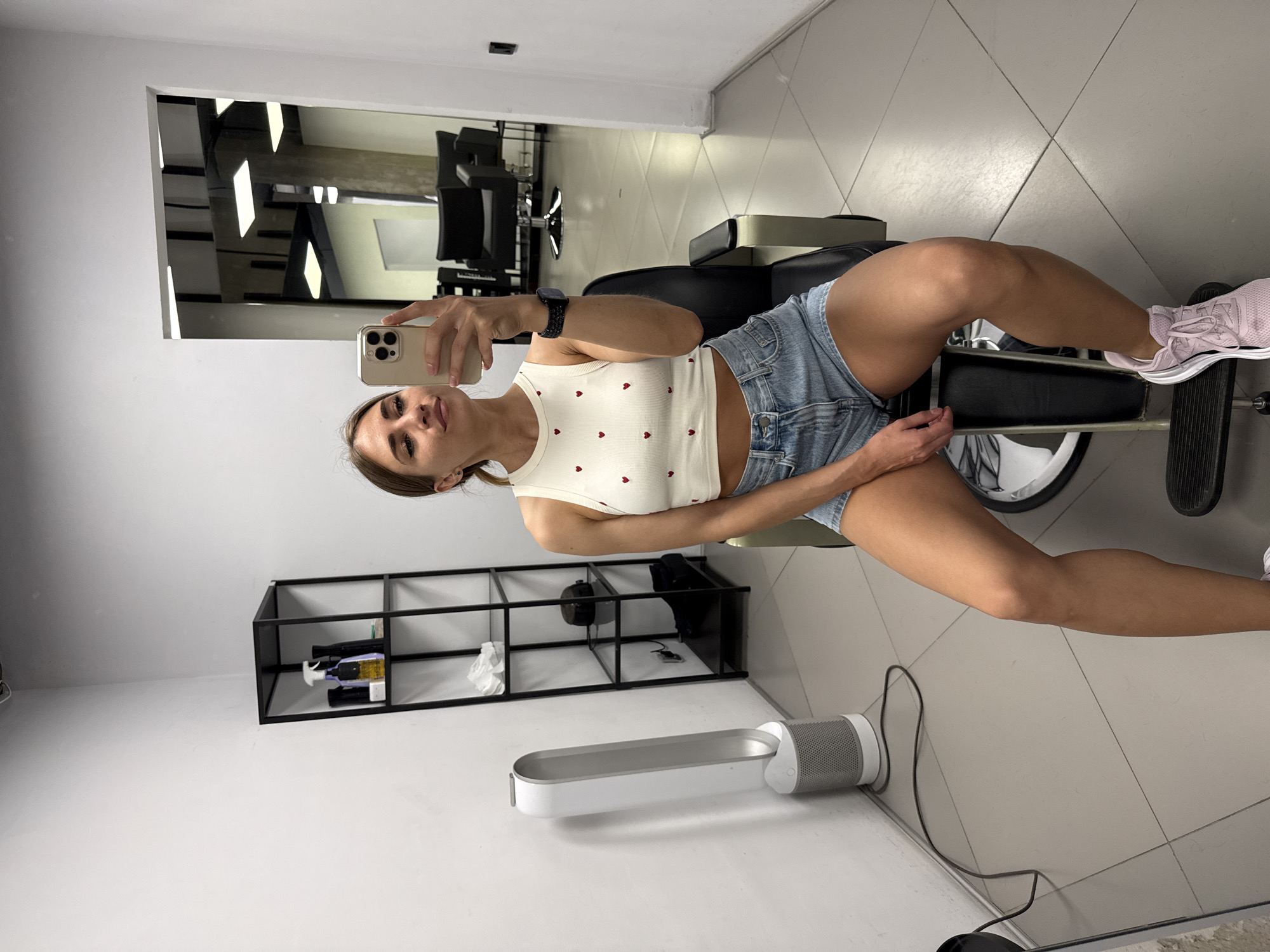 A young woman with long brown hair, wearing a white crop top with red hearts and denim shorts, sits on a black chair in a brightly lit room with white walls and tiled floors. She is taking a mirror selfie with a gold smartphone. To her left, a tall white tower fan stands near a black metal shelving unit holding various items, including bottles and bags. In the background, a doorway leads to another room with black salon chairs and large ceiling lights. The environment suggests a salon or barbershop. The scene is located in Saint Petersburg, Russia.