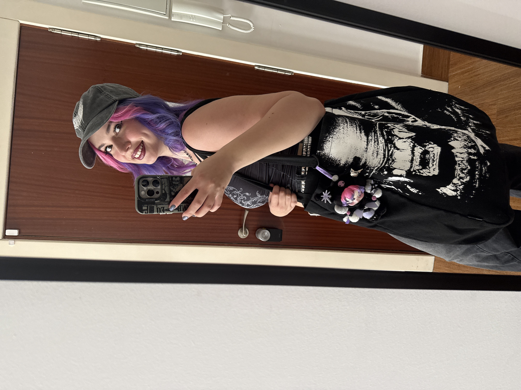 A young woman is taking a selfie in a mirror. She is wearing a grey baseball cap, a black tank top, a black skirt with a white graphic print, and grey pants. Her hair is dyed pink and purple. She is holding a smartphone in her left hand and smiling. The background shows a wooden door, a white wall, and a mirror frame. The location is impossible to determine from the given image.