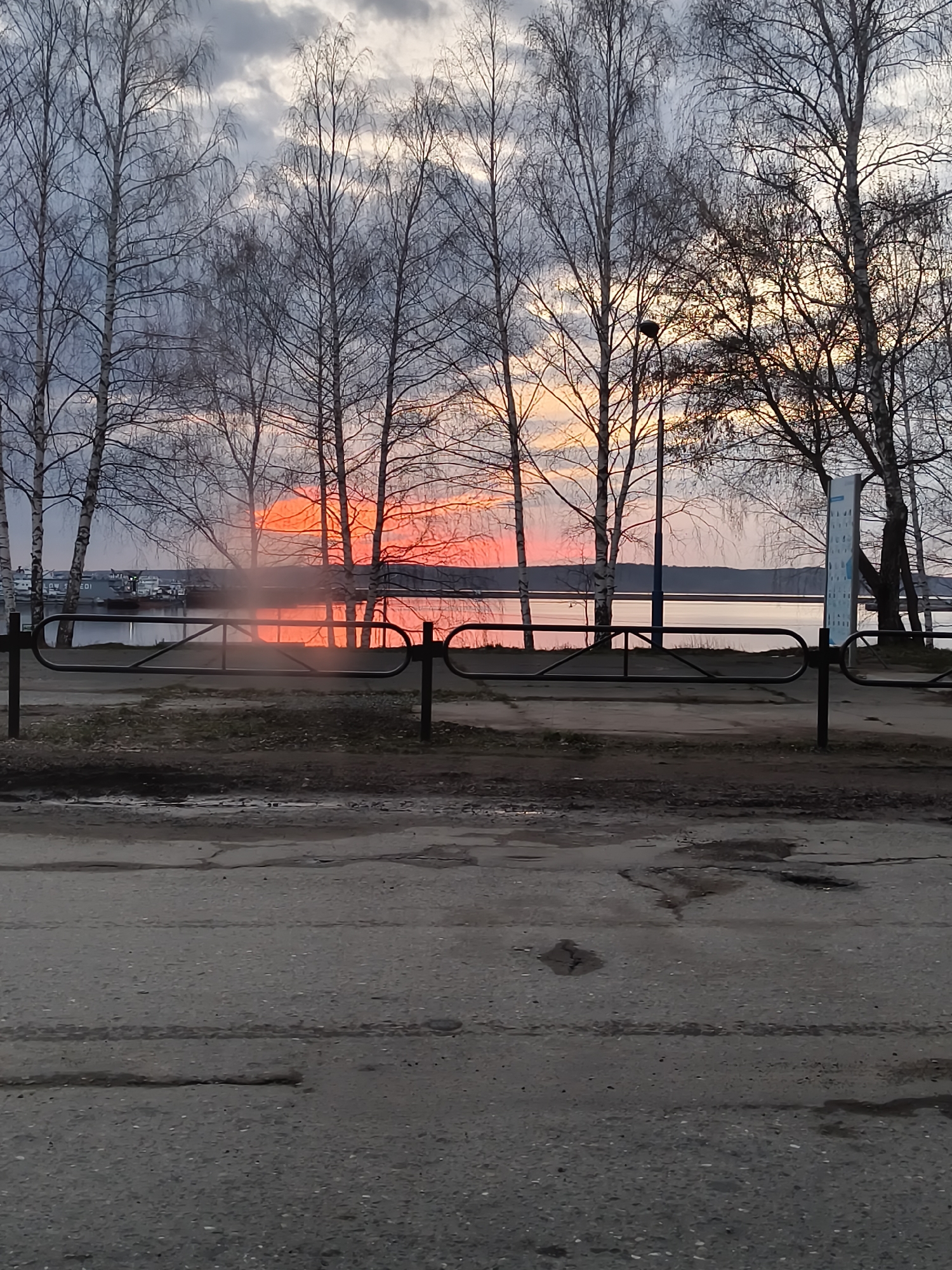 The image depicts a landscape at sunset. In the foreground, there is a grey asphalt road, slightly cracked and worn. A low black metal fence separates the road from a slightly elevated area of ground. Beyond the fence, there is a body of water, likely a lake or a wide river, reflecting the reddish-orange light of the setting sun. Several bare trees, typical of colder climates, line the water's edge. In the distance, there are some structures visible on the opposite side of the water. The sky is overcast with grey clouds, with the sunset colors breaking through the horizon. To the right, there is a lamppost and what appears to be a sign or informational board. No people are visible in the image. The location is Unknown, Unknown.