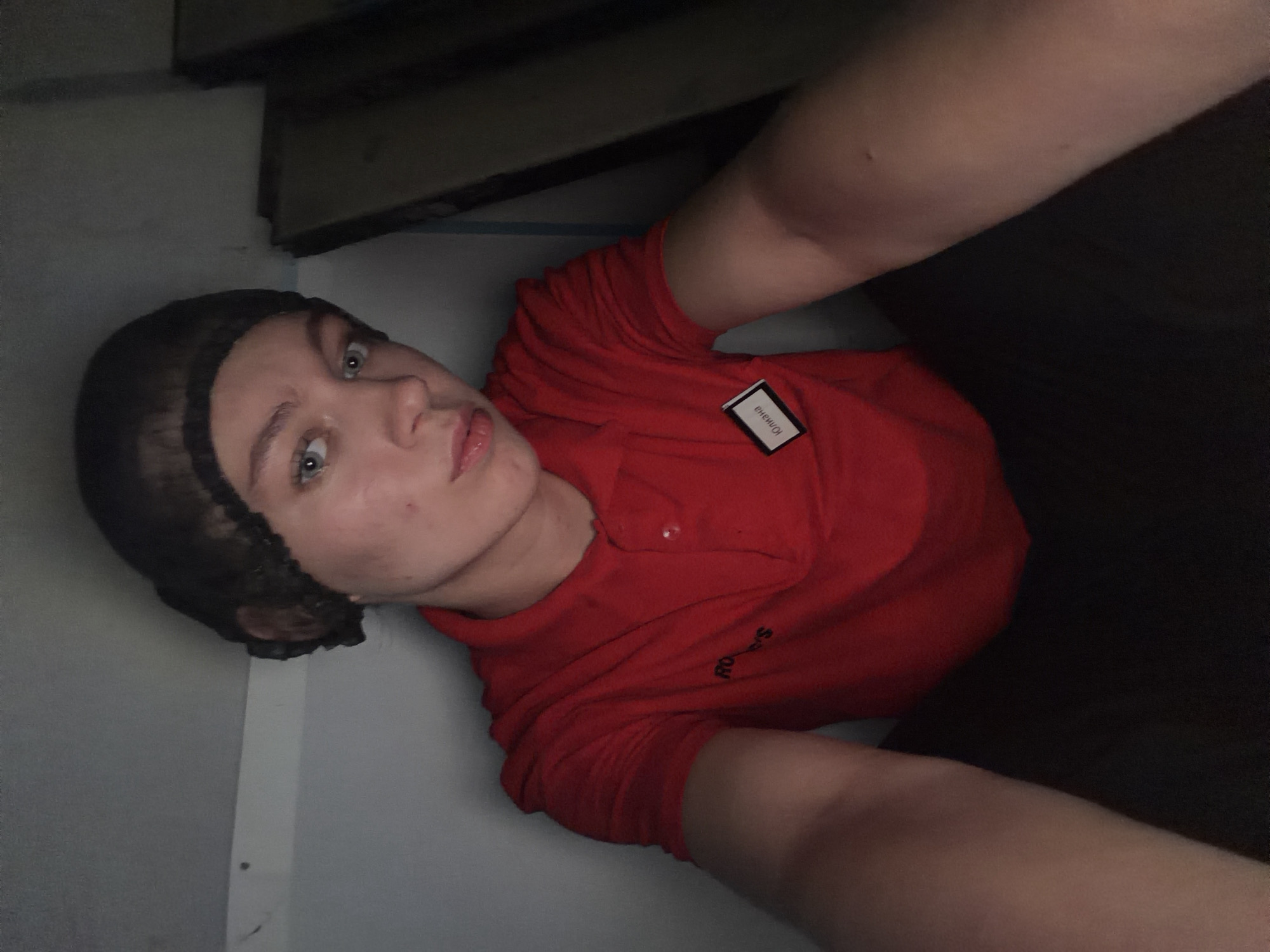 A young person wearing a red polo shirt with a black hair net sits in a dimly lit, confined space. The polo shirt has a small rectangular name tag on the left side. The person's face is partially illuminated, revealing light blue eyes and fair skin. The background appears to be a wall with some wooden structures or shelves. The overall impression is that of a utilitarian or backroom environment. The location is specified as Chaikovsky, Russia.