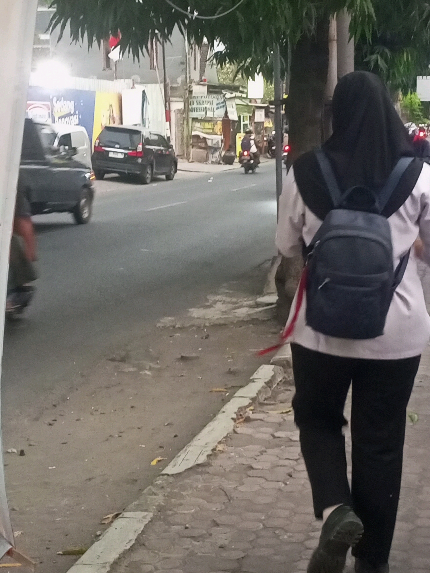 A medium shot captures a person wearing a black hijab and a light-colored long-sleeved shirt over black pants walking on a paved sidewalk. The person carries a dark-colored backpack. In the background, a black car is parked on the street, with other vehicles and buildings visible further down the road. The scene appears to be a street in Kediri City, Indonesia, indicated by the style of buildings and the presence of motorcycles, which are common in Indonesian street scenes. The sidewalk is constructed with hexagonal pavers and shows signs of wear and tear, with some debris scattered on it. The street itself is asphalt, with faded white lane markings. Trees line the side of the road, providing shade. The overall lighting suggests it is daytime.