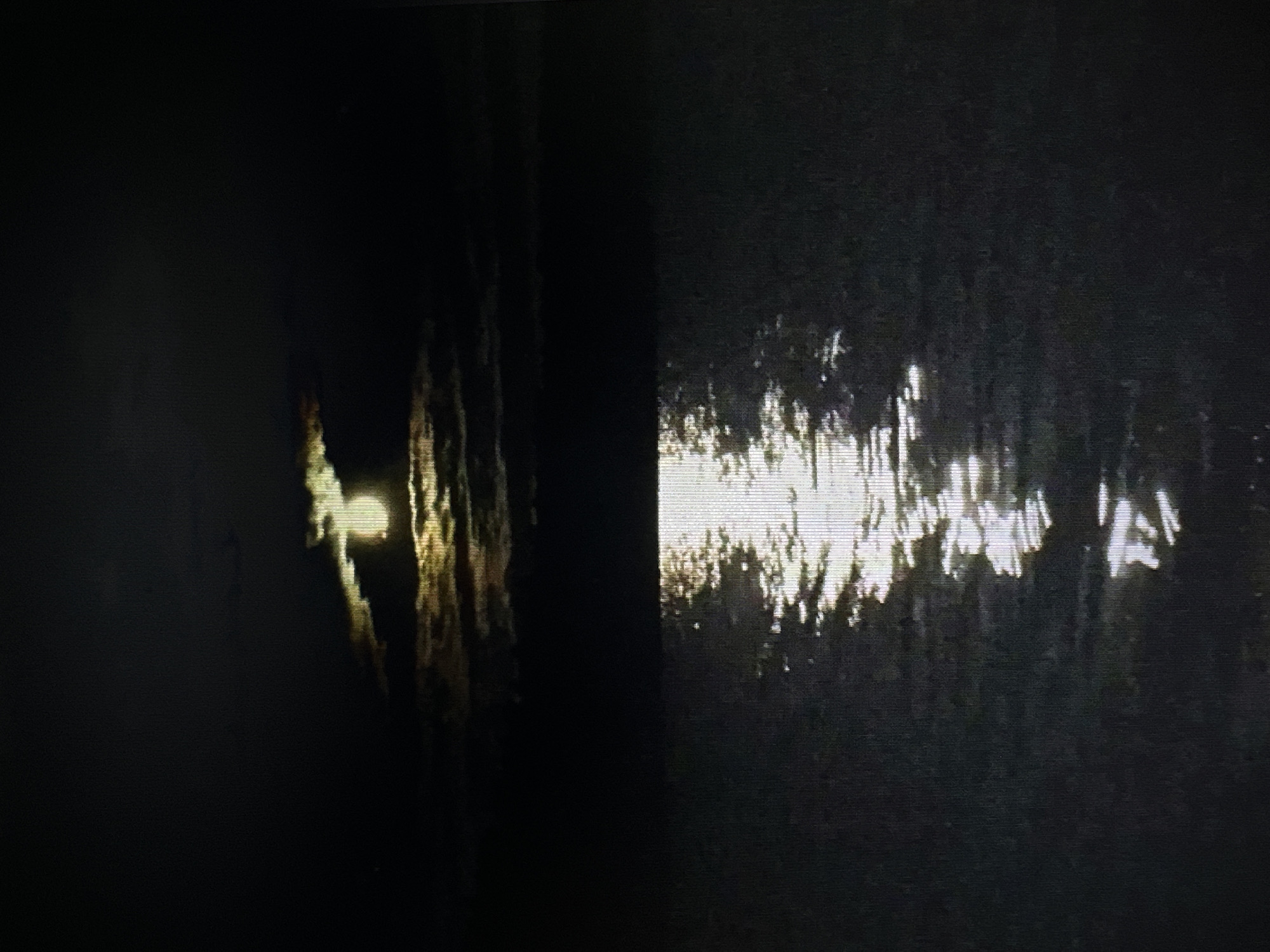 The image depicts a nighttime scene of the moon partially obscured by clouds, with its light reflecting on a body of water. The sky is dark with the bright moon creating a luminous glow. A prominent reflection of the moon, elongated and shimmering, stretches across the surface of the water. The water itself appears dark and rippling, with the moonlight creating a pathway of light. The environment is an outdoor, natural setting at night, with no visible human presence or identifiable city landmarks.
