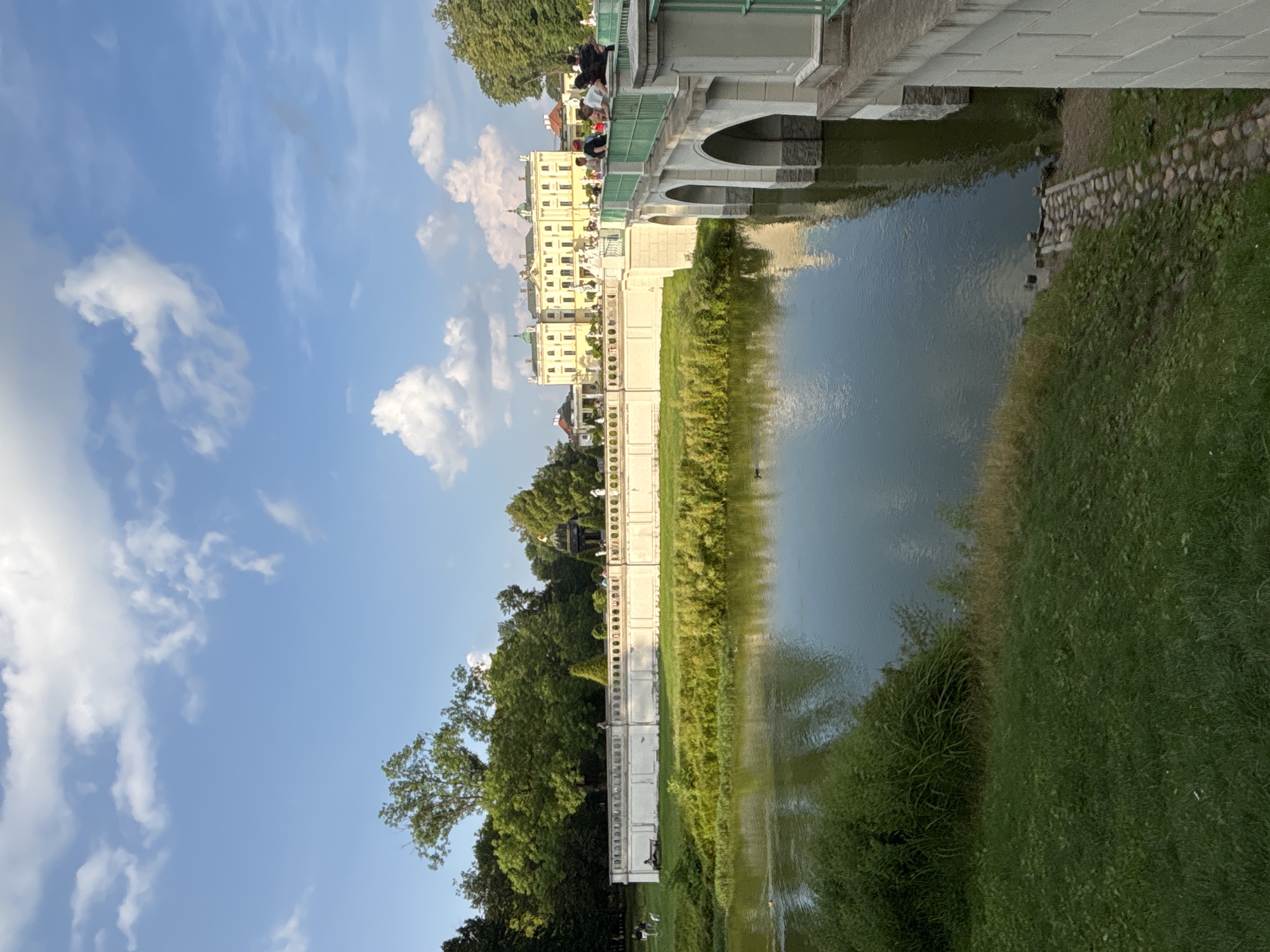 The image captures a scene of the Branicki Palace in Białystok, Poland, on a clear day with scattered white clouds in a blue sky. A calm body of water, likely a pond or river, occupies the foreground, with lush green grass and vegetation along its banks. In the midground, a white stone wall with decorative arches runs horizontally, separating the water from a well-maintained lawn. Beyond the wall, a grand, yellow-hued palace with a green roof and multiple wings is visible. A white bridge with arched supports crosses the water, leading towards the palace. Several people are present, with some standing on the bridge and others in the vicinity of the palace and the wall, appearing to be tourists or visitors. The overall atmosphere is serene and picturesque, showcasing the architectural beauty of the palace and its surrounding park.