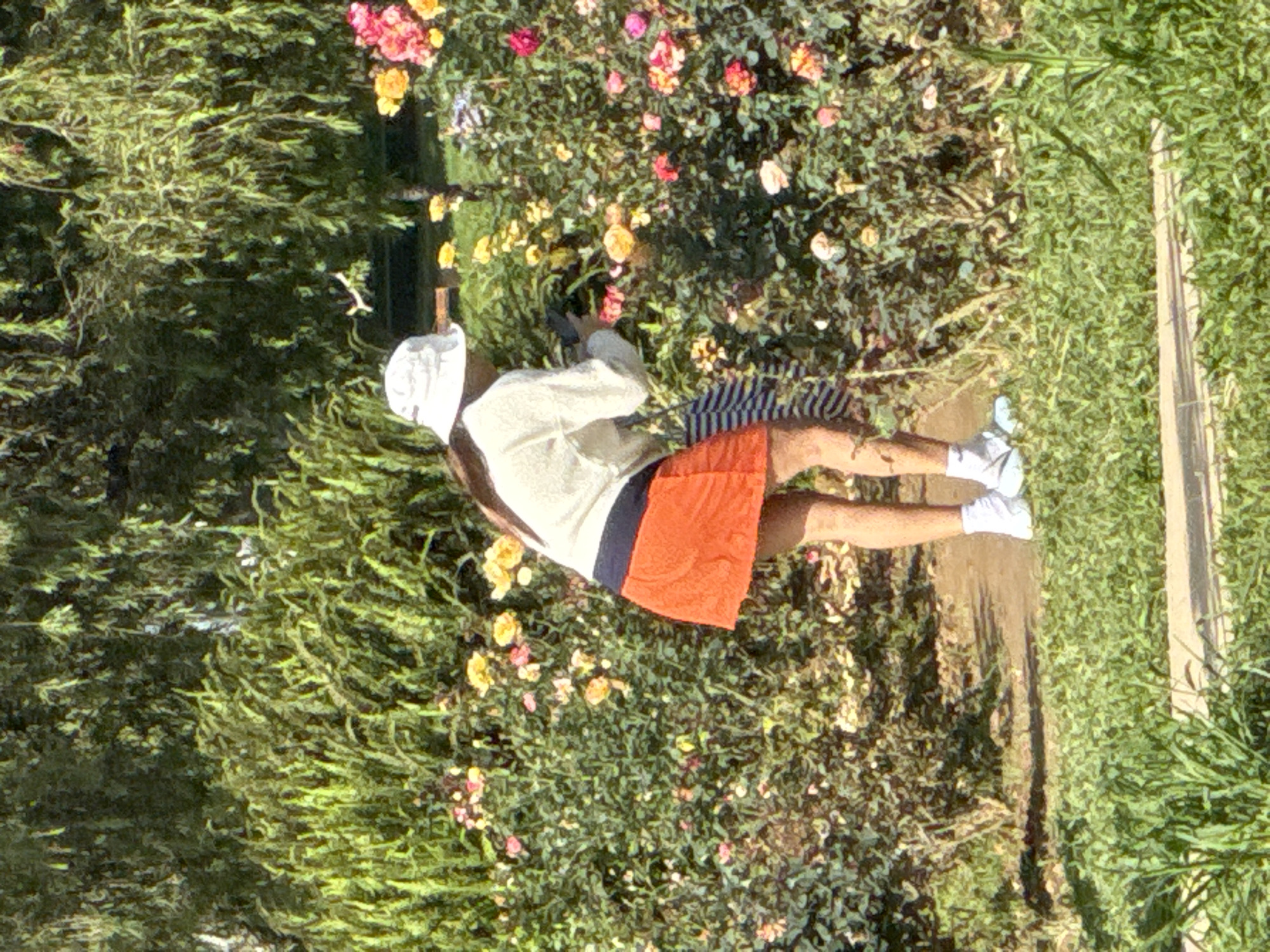 The image depicts an individual engaged in an activity within a garden or park setting in Phường Xuân Hương - Đà Lạt, Vietnam. The person, viewed from behind, is wearing a white bucket hat, a long-sleeved white top, an orange mini-skirt, white socks, and light blue/grey athletic shoes. They are bent forward, facing a dense bush of multi-colored roses, primarily yellow, pink, and orange. Both hands are holding a dark, rectangular object, consistent with a camera or smartphone, pointed towards the flowers, suggesting photography or close observation. A striped fabric item is visible near the person's left hip. The background consists of lush green foliage and trees, with bright green grass in the foreground and a visible dirt path where the individual stands.