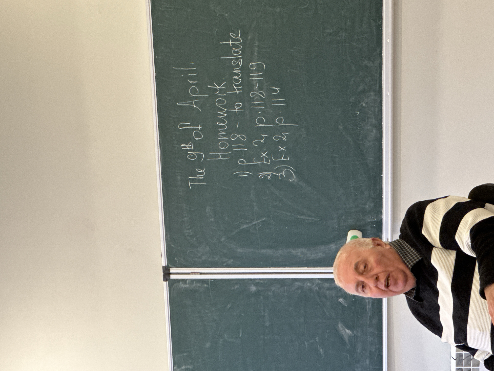 The image shows a man standing in front of a chalkboard with writing on it. The chalkboard says "The 9th of April. Homework. 1) p. 118 - to translate. 2) Ex 2, p. 118-119. 3) Ex 2, p. 114." The man is wearing a black and white striped sweater. The location is not mentioned in the image.
