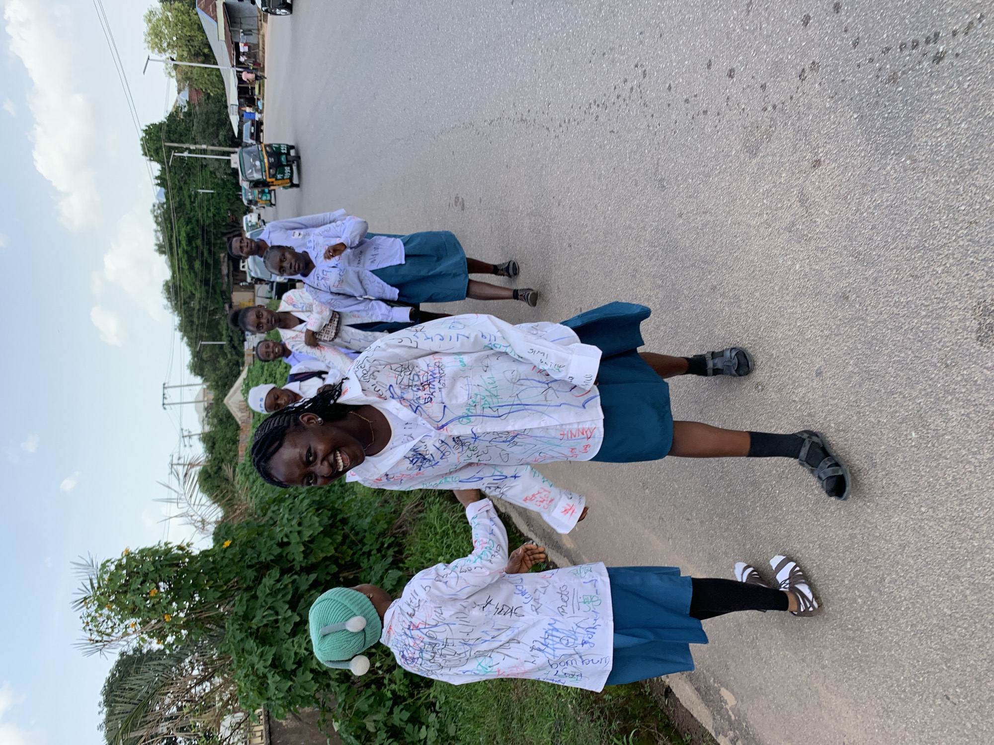 The image shows a group of young women, likely students, walking along a paved road lined with green vegetation and trees. They are dressed in white shirts, many of which are covered in colorful scribbled signatures and messages, possibly indicating a celebration or farewell. The women are wearing blue skirts and various footwear, including sandals and sneakers. The sky is visible in the background, with some clouds, suggesting it is daytime. There is a hint of a building with a corrugated roof and a street light in the distance, along with a small green and yellow auto-rickshaw, commonly known as a Keke Napep, on the road. One of the young women in the foreground is smiling broadly at the camera, conveying a sense of joy and excitement. Another woman in the foreground is wearing a green knitted hat with pom-poms. The overall scene appears to be in a somewhat rural or suburban area, likely in Jos, Nigeria, given the presence of the Keke Napep and the style of the buildings. The atmosphere is festive, suggested by the decorated shirts and the expressions of the individuals.