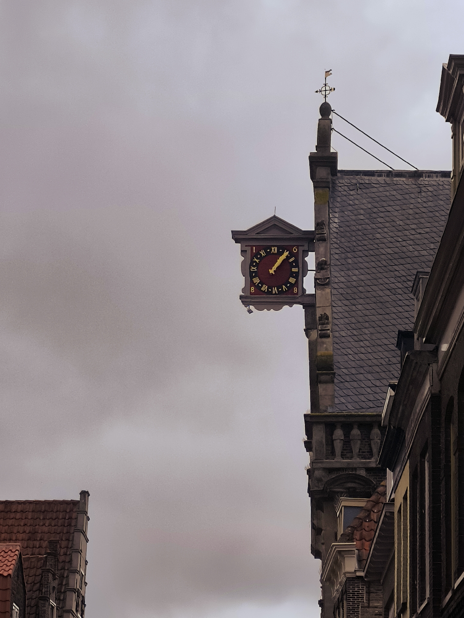 The image shows a street scene in Hoorn, Netherlands, featuring historic architecture. Prominently displayed on the side of a building is an ornate, red clock with Roman numerals and a yellow hand pointing to approximately 10:10. The clock is attached to a stone facade that extends upward to a slate-tiled roof. Above the roof, a weather vane with directional indicators is visible against a cloudy sky. To the left, another building with a red-tiled roof and stepped gables is partially visible. To the right, another building with dark brickwork and large windows frames the scene. The overall impression is of a historical urban environment under an overcast sky. There are no people or animals visible in the image.