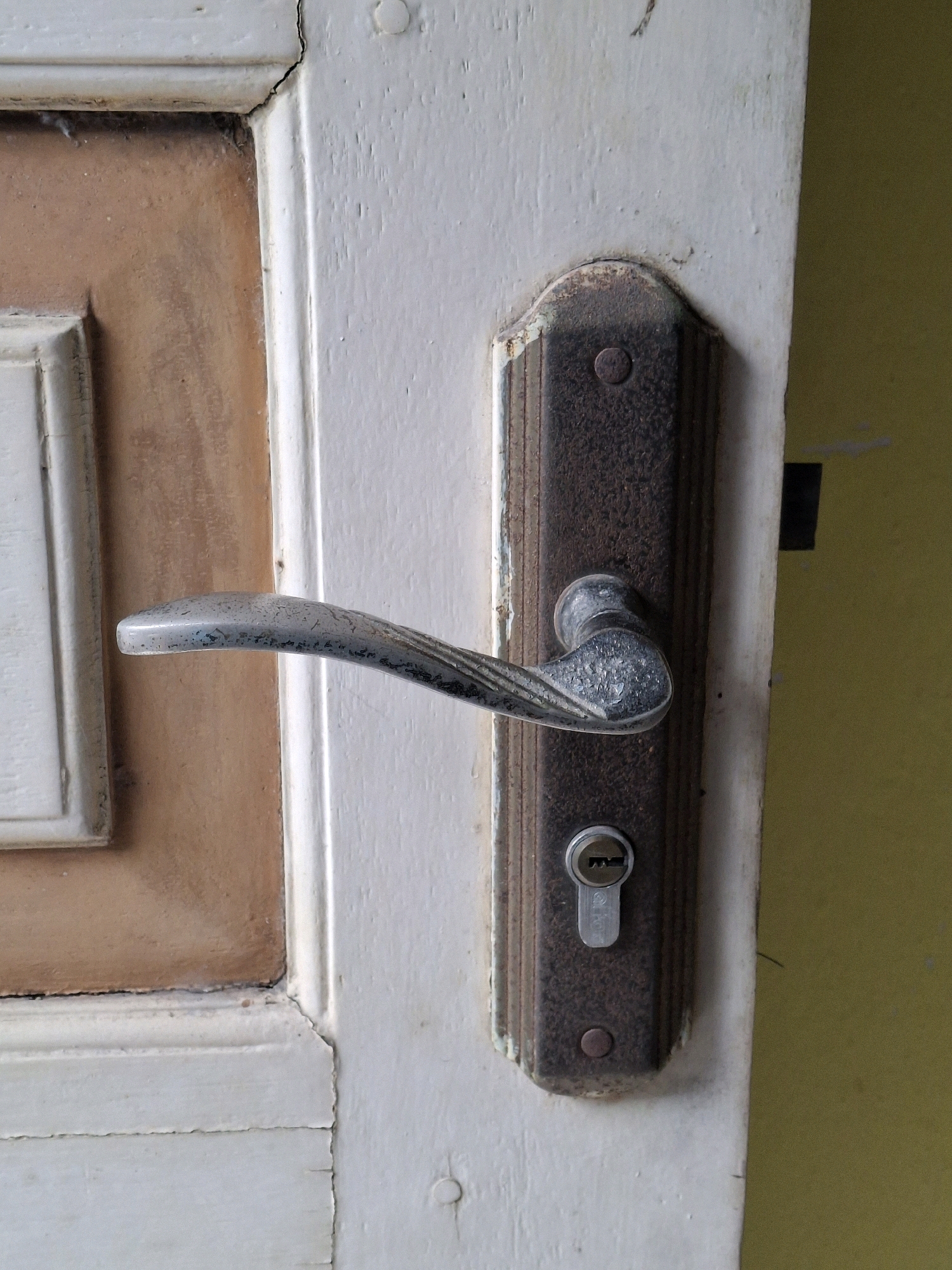 The image shows a close-up view of a door handle and lock mechanism on a weathered, white-painted door. The door has a panel with a light brown insert and a white frame. The door handle is made of silver-colored metal and is slightly curved. Below the handle is a dark brown metal plate with a keyhole. The plate is textured and appears to be slightly rusty around the edges. The door frame itself is also white and shows signs of age, with paint peeling and cracks. To the right of the door, a yellow wall is visible. The overall impression is of an older, somewhat worn entrance. The scene does not provide any specific indicators to pinpoint its exact location within Lumajang, Indonesia, other than it is a residential or building exterior.