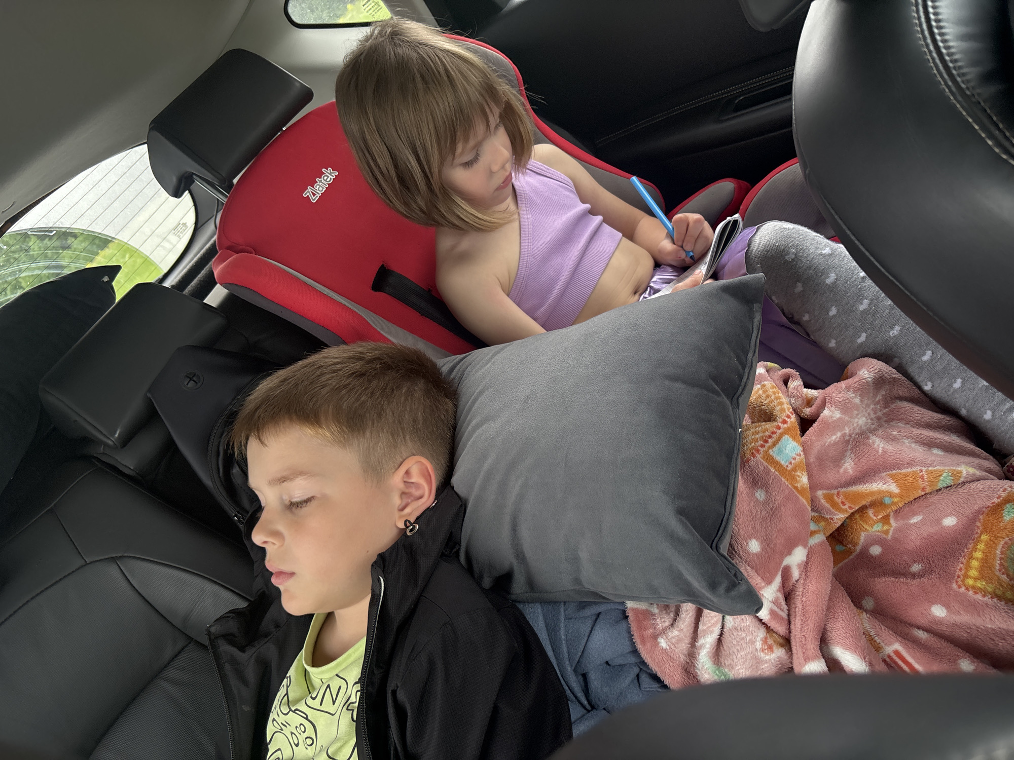 The image captures the interior of a vehicle in Nizhny Palnik, Russia, specifically the back seat area occupied by two children. On the left, a young boy with short brown hair is observed sleeping, positioned across black leather car seats. He wears a lime green t-shirt with a black design and a black zip-up jacket. His head rests on a grey pillow, and a small, dark, heart-shaped earring is visible on his left ear. Beneath him are several layered blankets, including a pink patterned one and a blue one. To the right, a young girl with shoulder-length light brown hair is seated in a red child safety seat, identified by the visible "Zlatek" branding. She is dressed in a purple one-shoulder crop top and matching purple pants, paired with grey patterned socks. The girl is actively engaged, holding a blue pen or marker and appearing to draw or write in an open book or notebook. Through the car's rear window, green foliage is discernible, indicating an outdoor, vegetated environment.