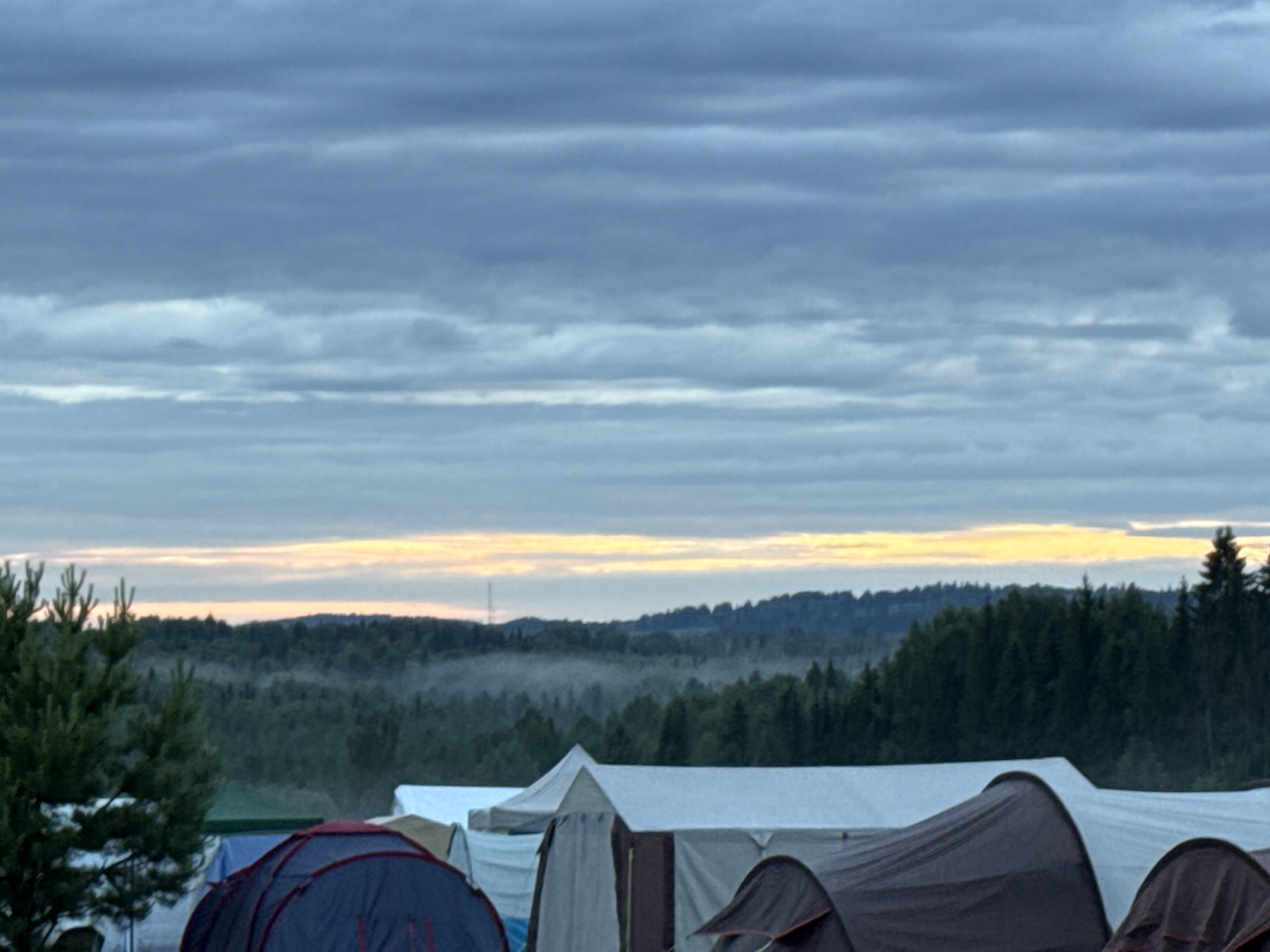This image captures an outdoor landscape scene at a campsite in Большая Кивара, Russia, characterized by subdued light, likely during dawn or dusk. The upper two-thirds of the frame are dominated by an overcast sky, presenting layers of grey and muted blue clouds in horizontal formations. A distinct, narrow band of yellow-orange light stretches along the horizon, separating the sky from the distant terrain. Below this light, a vast, dense forest covers rolling hills, with a visible layer of low-lying mist or fog settled within a valley in the mid-left to center background. A slender, dark, vertical structure, possibly a communication tower, is faintly discernible rising above the distant treeline to the left of the central axis. In the foreground, several camping tents are visible. A dark blue and red domed tent occupies the left foreground, partially obscured. To its right and extending towards the center and right foreground are multiple other tents, appearing in shades of white, off-white, brown, and dark grey. A prominent evergreen tree with dense green foliage stands in the far left foreground, partially obscuring the lower midground. No human figures are present or discernable within the depicted scene.