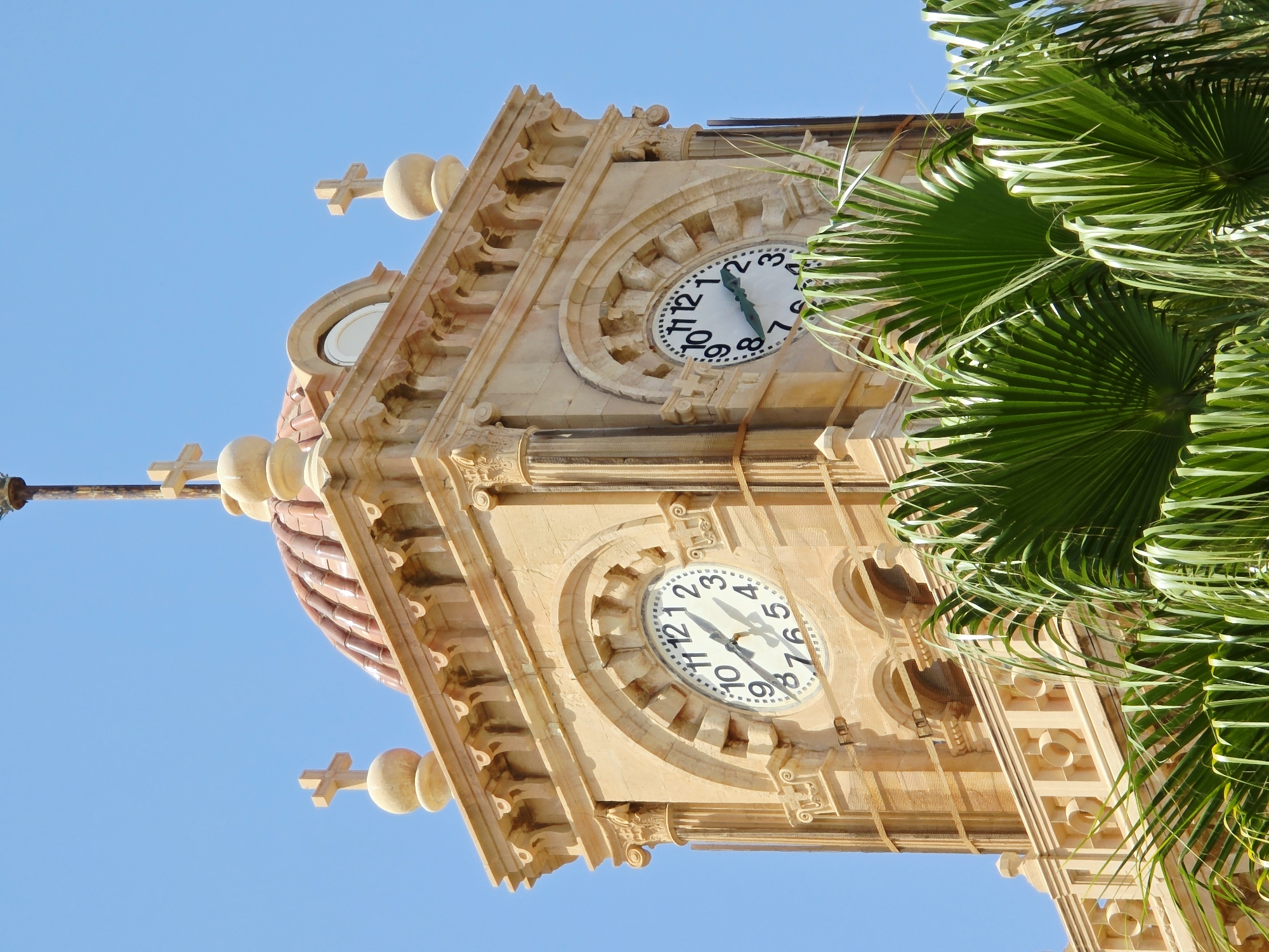 An upward shot of a tan-colored clock tower with a copper dome under a clear blue sky. Two large, round clocks are visible on the tower's sides, each showing a different time. The clock on the left indicates approximately 10:47, while the one on the right shows about 10:49. The tower is adorned with architectural details, including arches, columns, and decorative moldings. Atop the tower are several small crosses. In the foreground, vibrant green palm fronds partially obscure the lower section of the structure. The architectural style suggests it may be a significant historical or religious building. This is the Venetian Loggia in Heraklion, Crete, Greece.