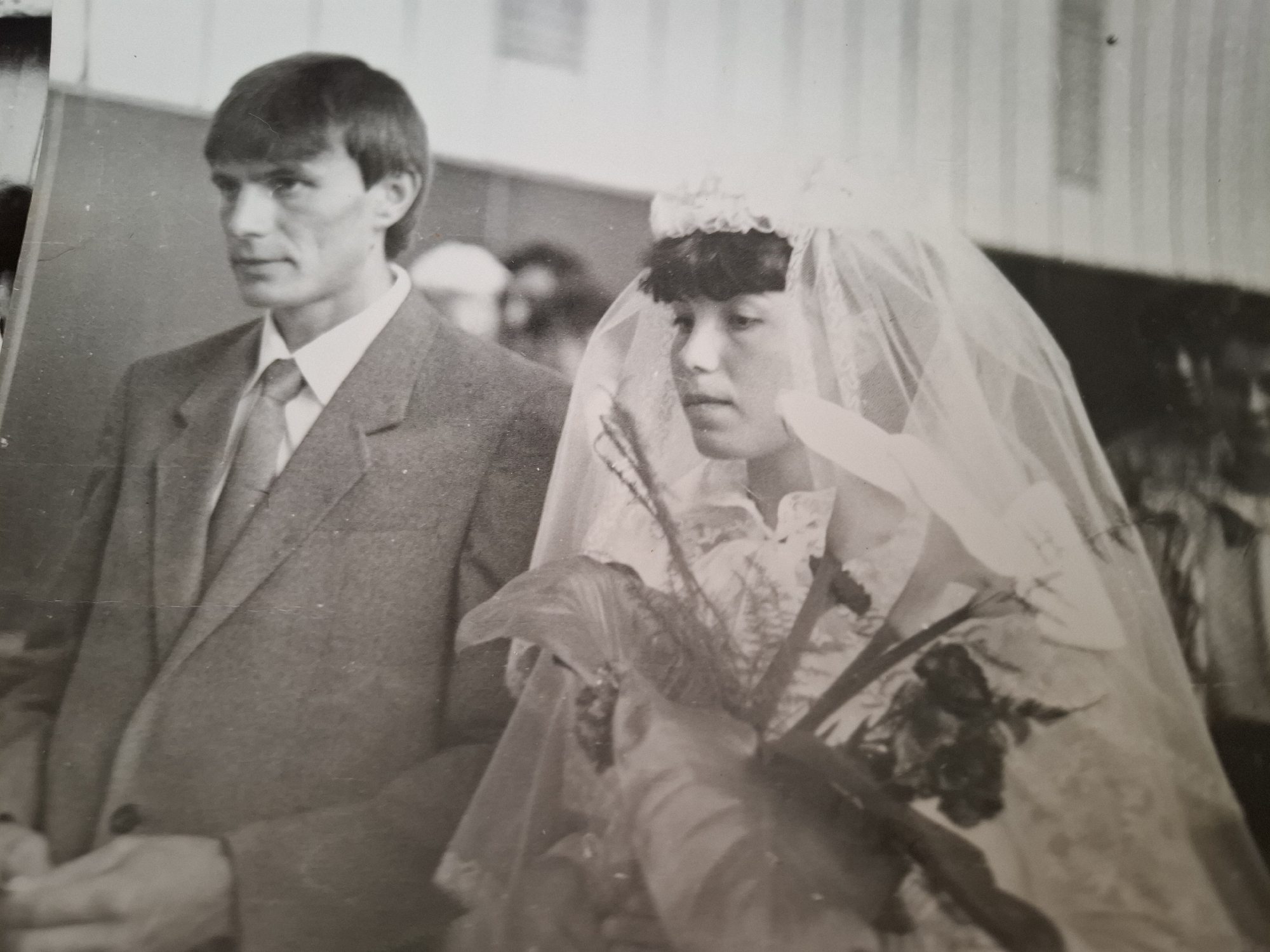 A black and white photograph depicts a wedding scene in the city of Амзя, Russia. A man in a suit and tie stands next to a woman in a wedding dress and veil. The woman is holding a bouquet of flowers. Other people are present in the background.