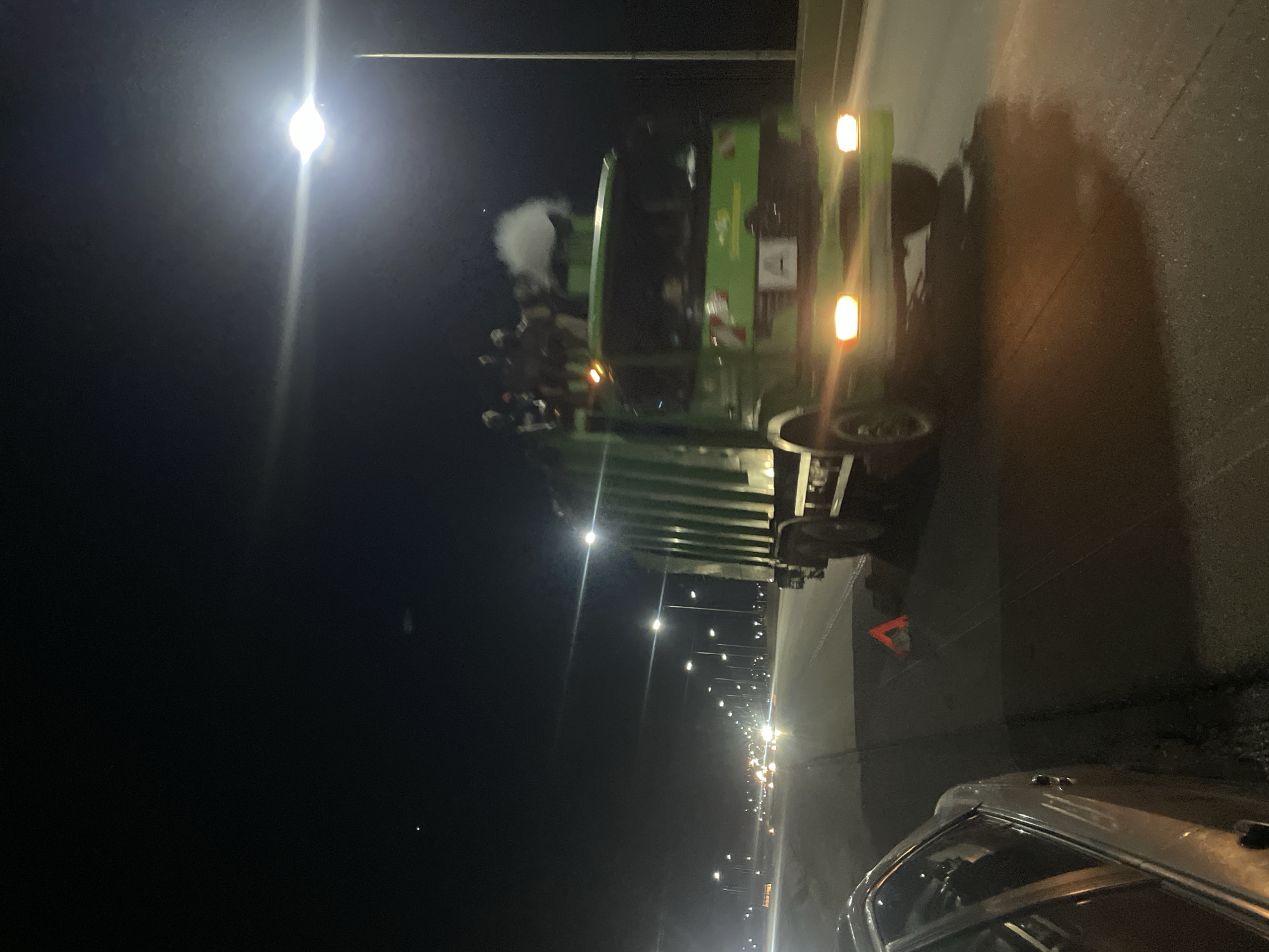 At night on a highway, a green garbage truck with people riding on top drives on the right side of the road. In the foreground on the left, a silver car is parked on the shoulder. An orange triangular warning sign is placed on the road behind the car. Streetlights illuminate the highway, with some fog or mist visible in the air. The scene appears to be within the vicinity of Mopol 44 Barracks Kuje, Nigeria, a location commonly associated with military police and possibly serving as a transportation hub or residential area.