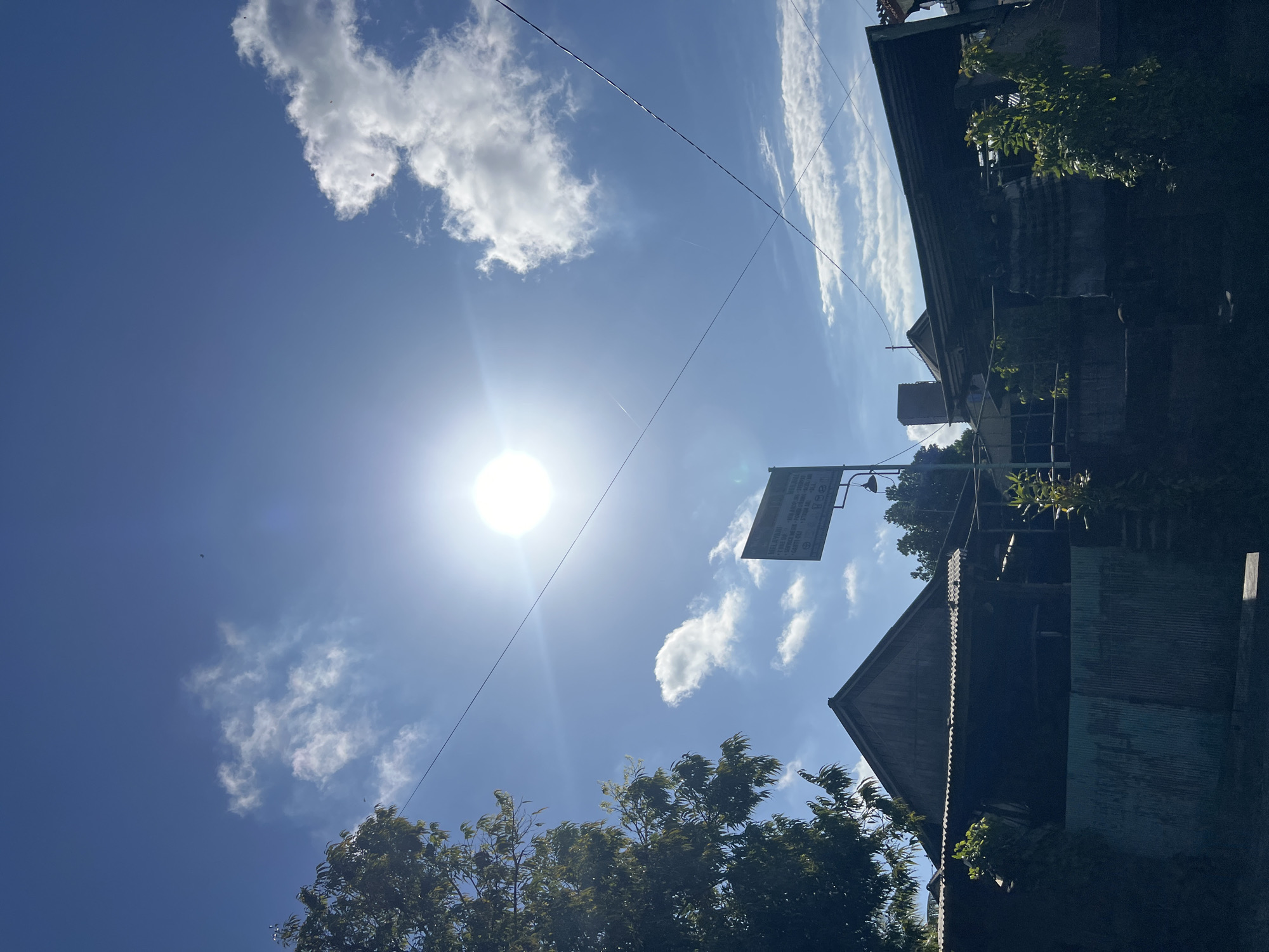The image depicts a bright, sunny day in Sukawati, Indonesia, with the sun prominently positioned in the upper center of the frame, casting a lens flare. The sky is a vibrant blue, dotted with scattered white clouds. In the foreground, a cluster of traditional Indonesian buildings with corrugated metal roofs and wooden structures are visible. Trees with lush green foliage frame the left side of the scene, and a metal fence is present in front of one of the buildings. A signpost with text is also visible in the middle ground, and electrical wires crisscross the sky. The overall impression is of a typical, warm day in a rural or semi-rural area of Sukawati.