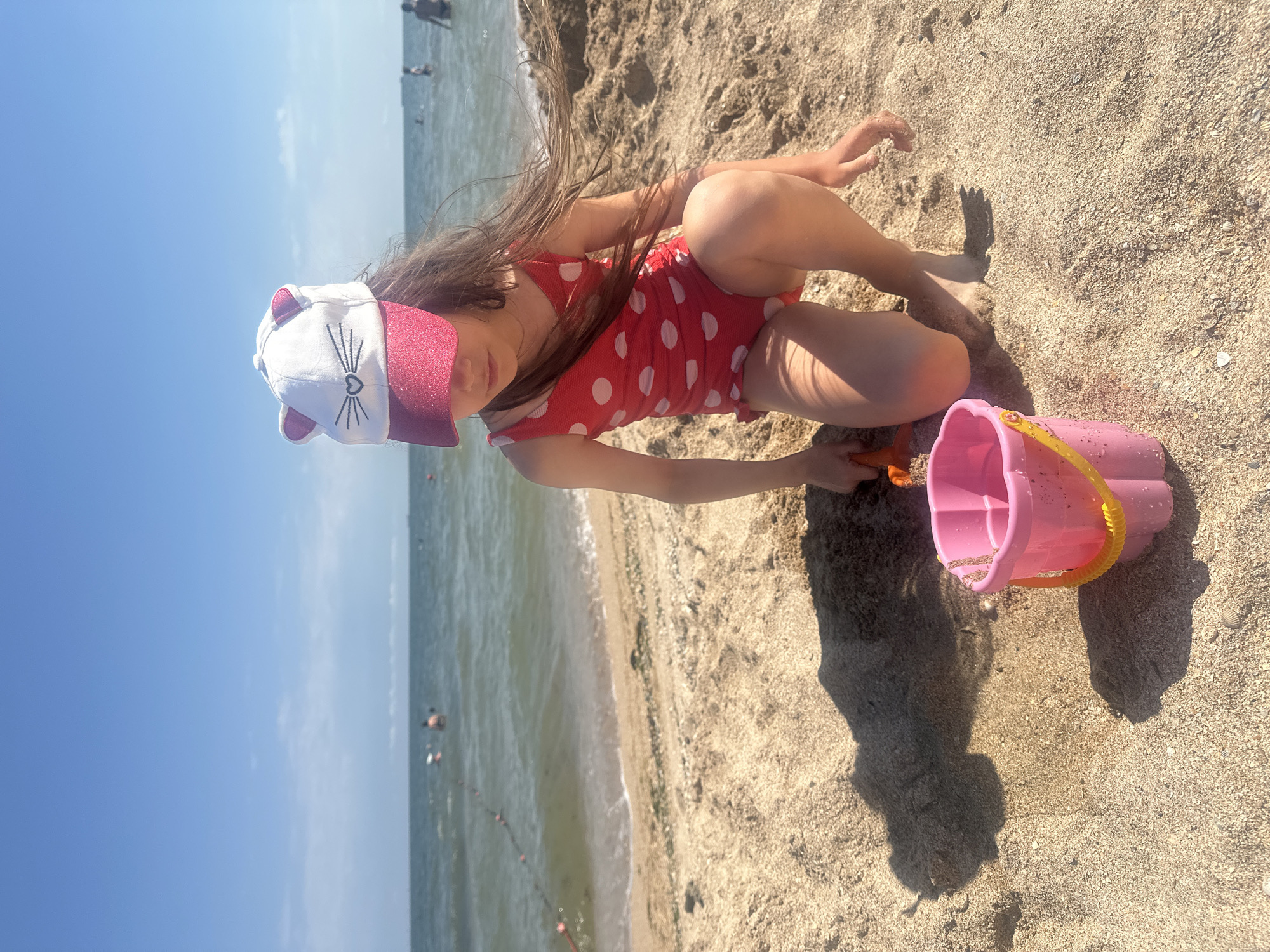 A young girl wearing a red polka dot swimsuit and a white cap with cat ears and a glittery brim is playing in the sand on the beach in Golubitskaya, Russia. She is crouching down, holding a small orange shovel, and looking at a pink bucket with a yellow handle. The sand is light brown and speckled with shells. In the background, the sea is visible with a few people swimming and a rope with buoys stretching across the water. The sky is clear blue.