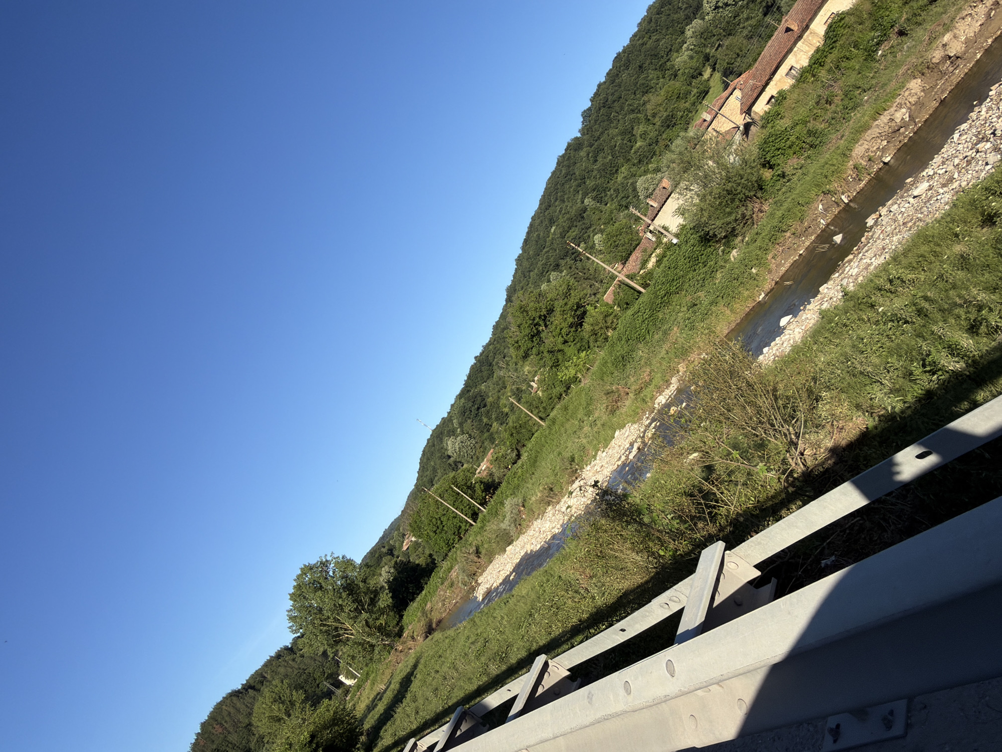 The image is a landscape photograph taken from a slightly elevated position, possibly a bridge or overpass, looking down onto a rural scene in the city of Veselinovo, Bulgaria. The photograph is tilted, with the sky occupying the upper right portion of the frame and the ground filling the lower left.
In the foreground, a metal guardrail runs diagonally from the bottom left corner upwards and to the right. It is constructed of thick, grey metal bars with visible bolts and rivets. The guardrail casts a distinct shadow across the grassy embankment to its right.
Beyond the guardrail, a shallow, rocky stream flows from the background towards the foreground. The stream bed is composed of various sizes of grey and brown stones. The banks of the stream are covered in dense green grass and interspersed with small shrubs and weeds.
The terrain slopes upwards from the stream, forming a grassy hill. Scattered along the hillside are several poles with electrical insulators, suggesting utility infrastructure. Further up the slope, there are clusters of trees and patches of green vegetation.
In the middle ground and background, several buildings with terracotta-tiled roofs are visible, nestled amongst the trees and on the hillside. These appear to be traditional rural homes. The architecture suggests an older, possibly rustic style.
The entire scene is bathed in bright, natural sunlight, indicating it was taken during the daytime. The sky is a clear, vibrant blue, with no clouds visible. The overall impression is one of a serene, sunny day in a hilly, rural area.