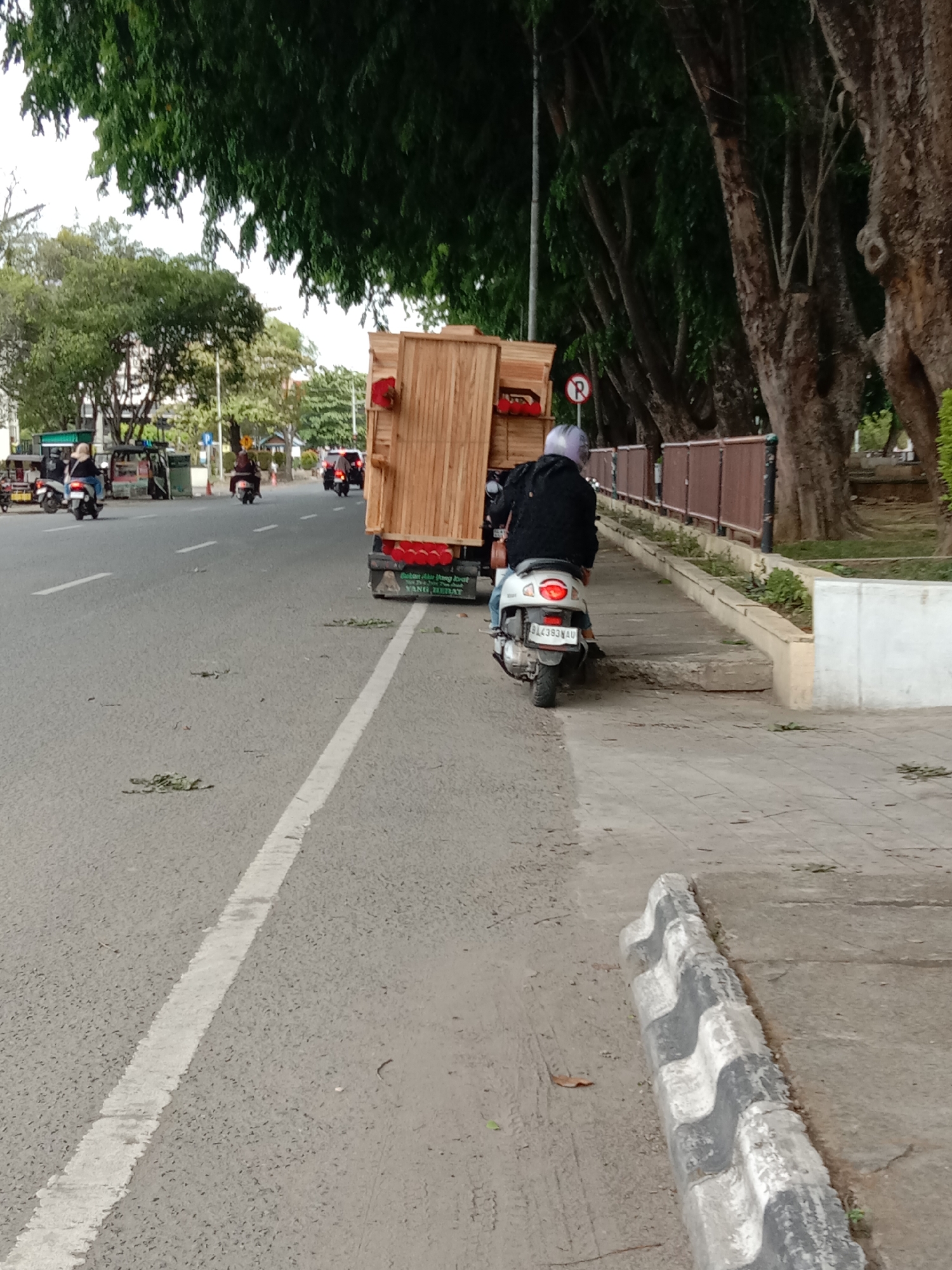 A street scene shows a person riding a scooter with a large wooden structure on the back. The scooter is on the side of the road near a sidewalk. Other scooters and vehicles are visible further down the road. The area is lined with trees on one side, and there are road markings on the asphalt. Fallen leaves or small branches are scattered on the road. A black and white striped barrier is in the foreground. The location appears to be an urban environment.