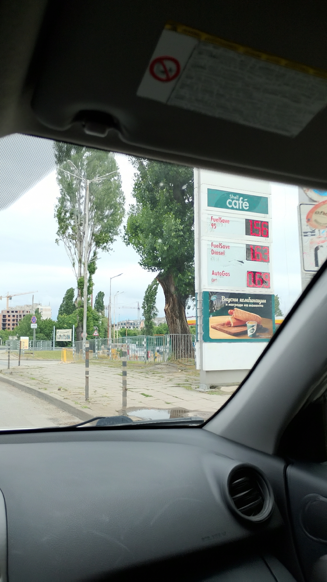 The image, taken from inside a vehicle in Sofia, Bulgaria, captures a view of a Shell gas station. A large price board is prominently displayed, showing fuel prices for FuelSave 95 (1.96), FuelSave Diesel (1.63), and AutoGas (1.63). Below the fuel prices, an advertisement for "Вкусни комбинации" (delicious combinations) featuring a hot dog and coffee is visible. The gas station is surrounded by greenery, with tall trees and a partly constructed building in the background. The sky is overcast. The interior of the car is partially visible, including the dashboard and a sun visor with a warning sticker.