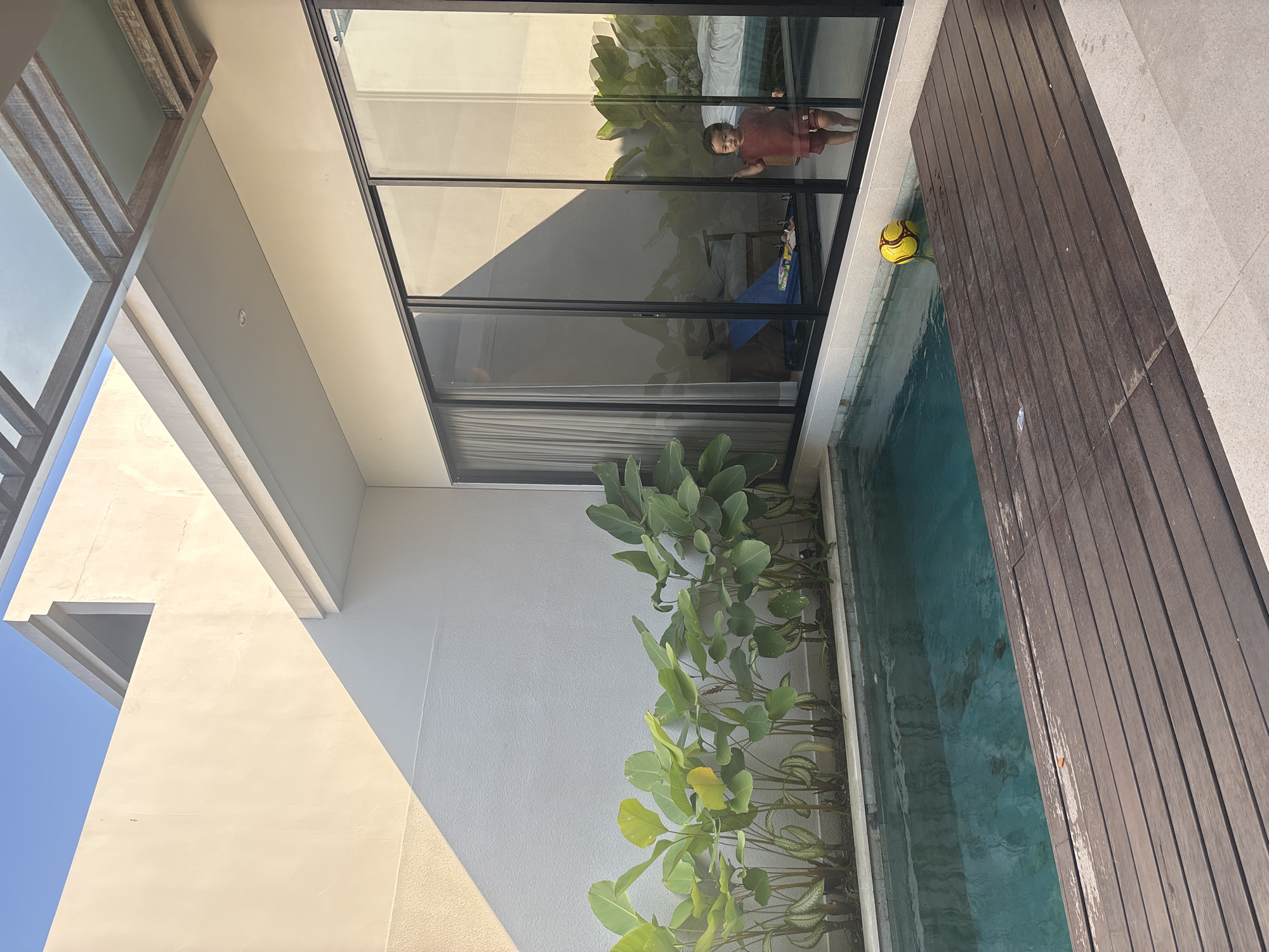 A young child stands near a large glass sliding door in a modern villa courtyard in North Kuta, Indonesia. The child, wearing a maroon t-shirt and shorts, is reflected in the glass. Outside, a small, turquoise swimming pool is bordered by a wooden deck and lush green plants. A yellow and black soccer ball floats in the water near the edge of the deck. The courtyard features a white stucco wall, partially covered in sunlight and shadow, and a covered patio area with a wooden beam ceiling. The background inside the villa appears to be a bedroom with a bed and furniture.