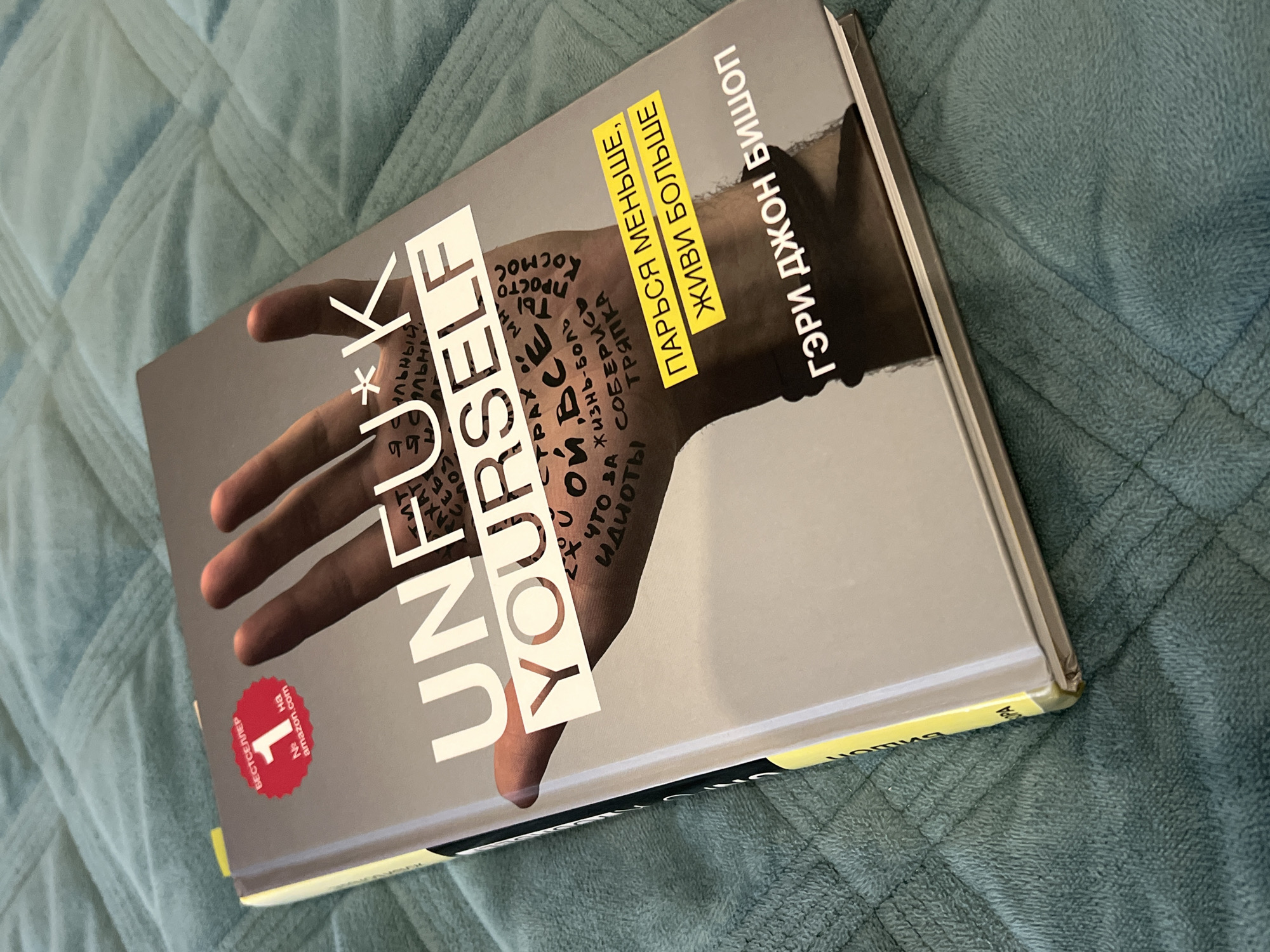 The image shows a Russian translation of the book "Unfu*k Yourself" by Gary John Bishop. The book is laying on a blue quilted surface. The cover is gray and features a hand with words written on it. The book has a yellow binding. A red sticker on the bottom left corner indicates it was a bestseller on Amazon.com. The text on the cover is in both English and Russian. There is no indication of the location being Chaikovsky, Russia.