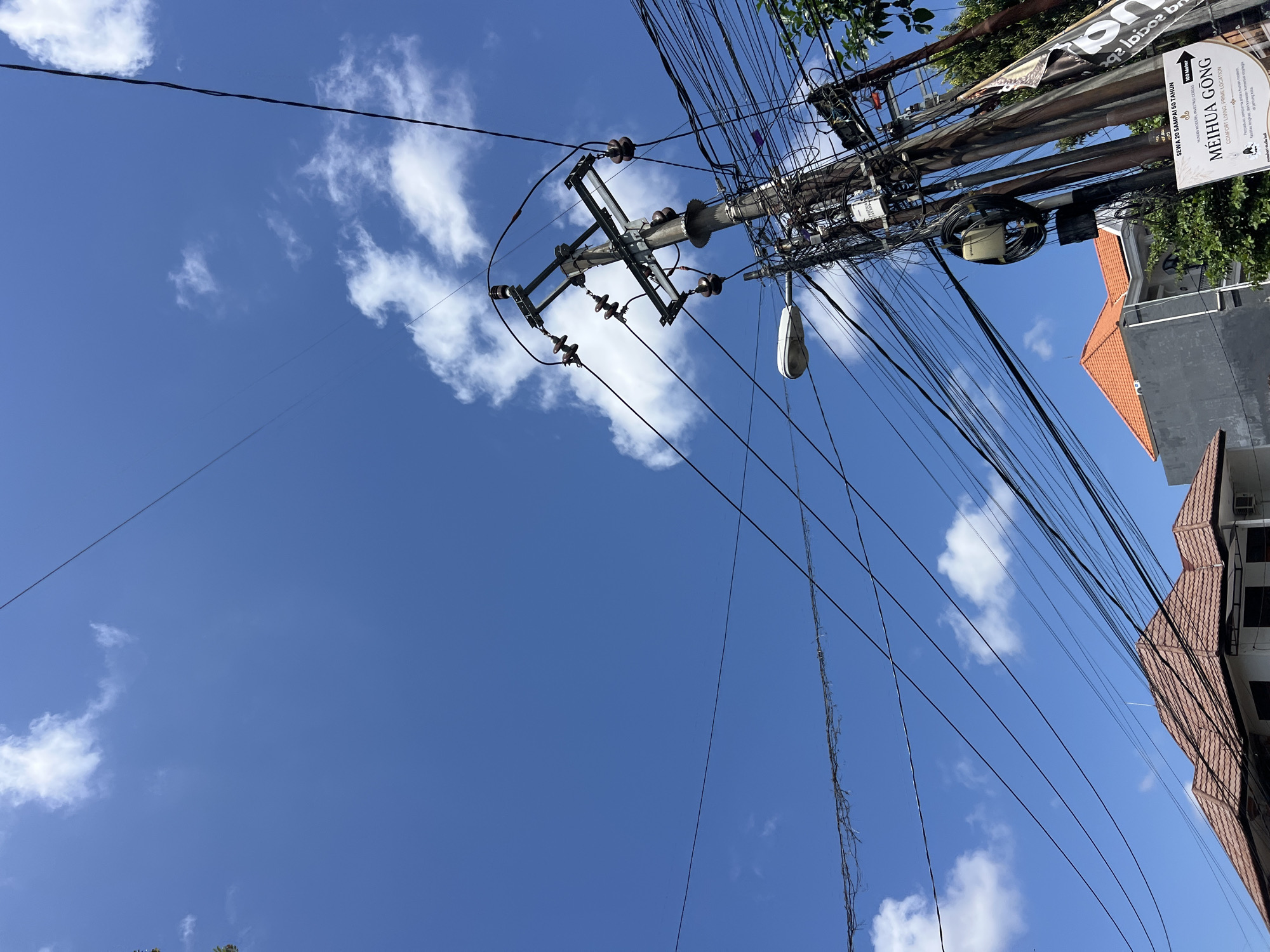 An eye-level, slightly low-angle outdoor shot captures a utility pole with numerous tangled wires against a bright blue sky dotted with white clouds. A street lamp is attached to the pole. In the lower left and center, the terracotta tiled roof of a building is visible. To the right of the pole, a white sign with black text is partially obscured by foliage. The scene appears to be a street in Denpasar, Indonesia, as indicated by the text on the sign which reads "SEWA 20 SAMPAI 50 TAHUN," a phrase commonly seen on property rental advertisements in Indonesia, and mentions "MEIHUA GONG" and "COMFORT LIVING PRIME LOCATION." The overall impression is of an urban or semi-urban street scene on a clear day.