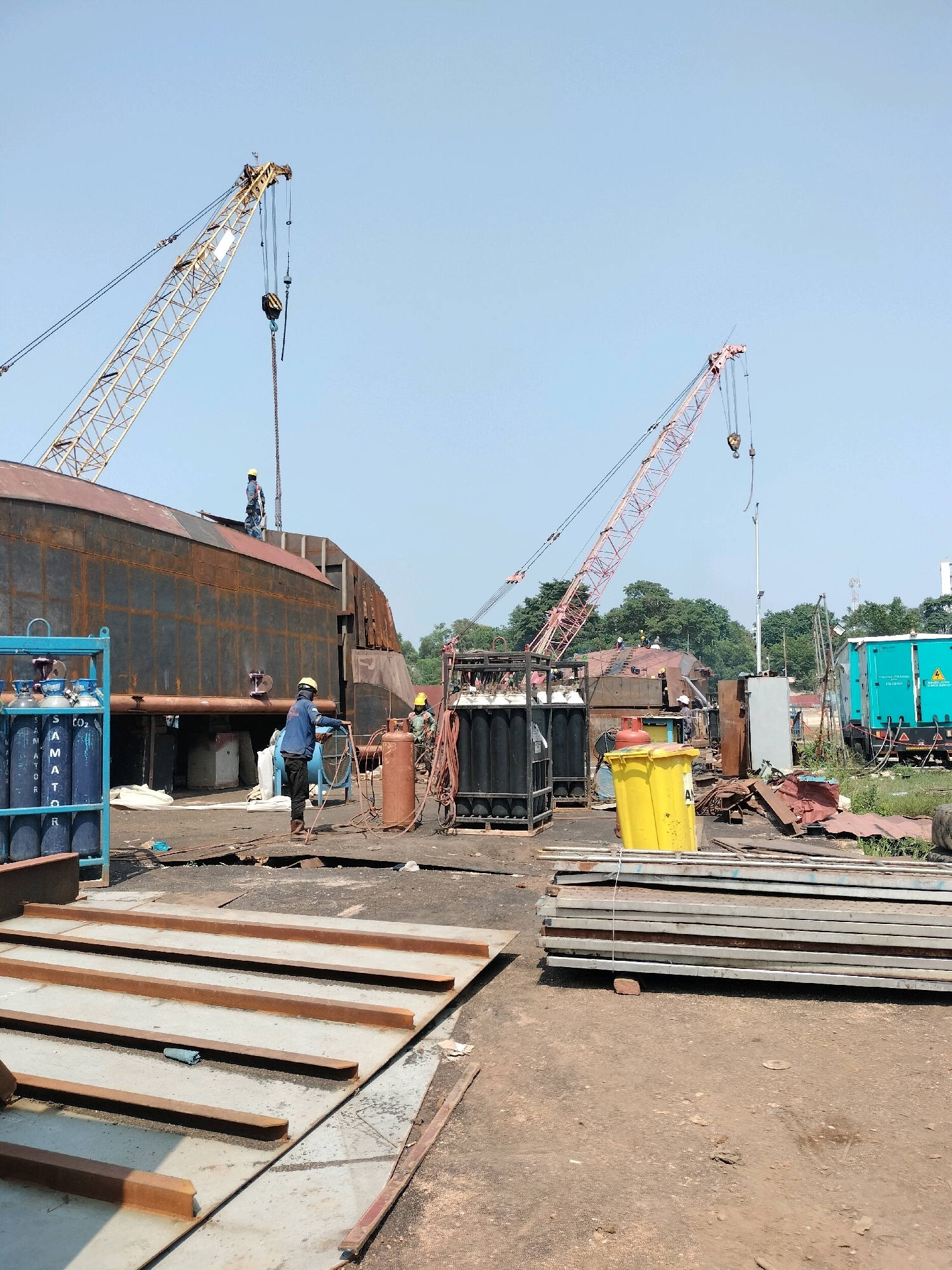 The image depicts an outdoor industrial scene in Batam, Indonesia, likely a shipyard or construction site. Two large cranes, one yellow and one pink, dominate the upper portion of the frame, with their booms extended. In the midground, a large, dark, rusted metal structure, possibly a ship hull or large industrial component, is under construction. Several workers in hard hats are visible, some near the structure and others in the foreground. One worker is operating welding equipment near a stack of oxygen and other gas cylinders. In the foreground, stacks of flat metal sheets and beams are laid out on the ground, along with a blue cart holding several gas cylinders labeled "SAMATOR CO2". A bright yellow bin is also present. The environment appears to be dusty and industrial, with some greenery and buildings visible in the background. The sky is clear and blue.