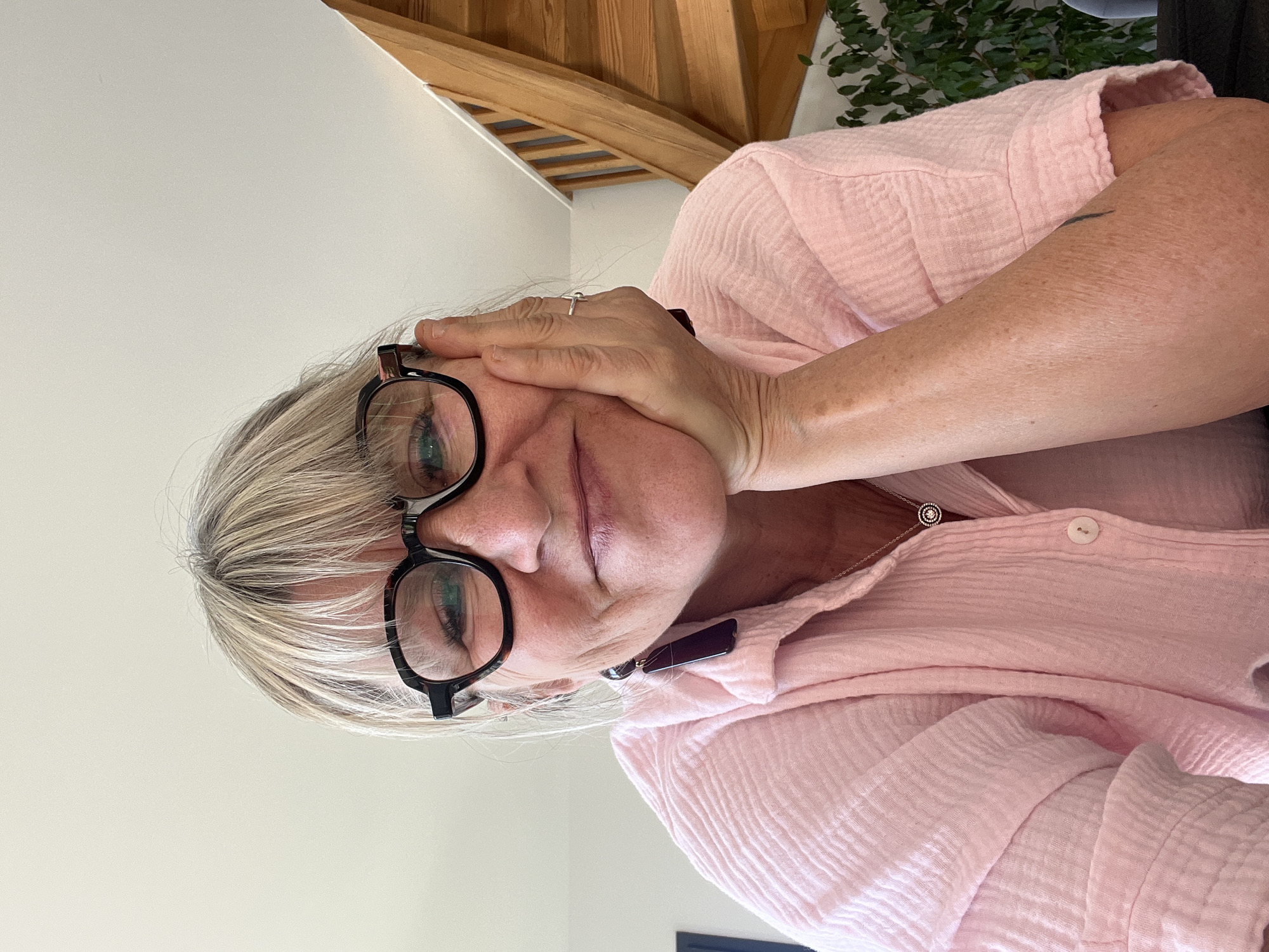 A woman with blonde hair and glasses rests her head in her hand, wearing a pink shirt. She is indoors with a wooden beam structure visible above. The location, Chambœuf, France, is not discernible from the image alone.