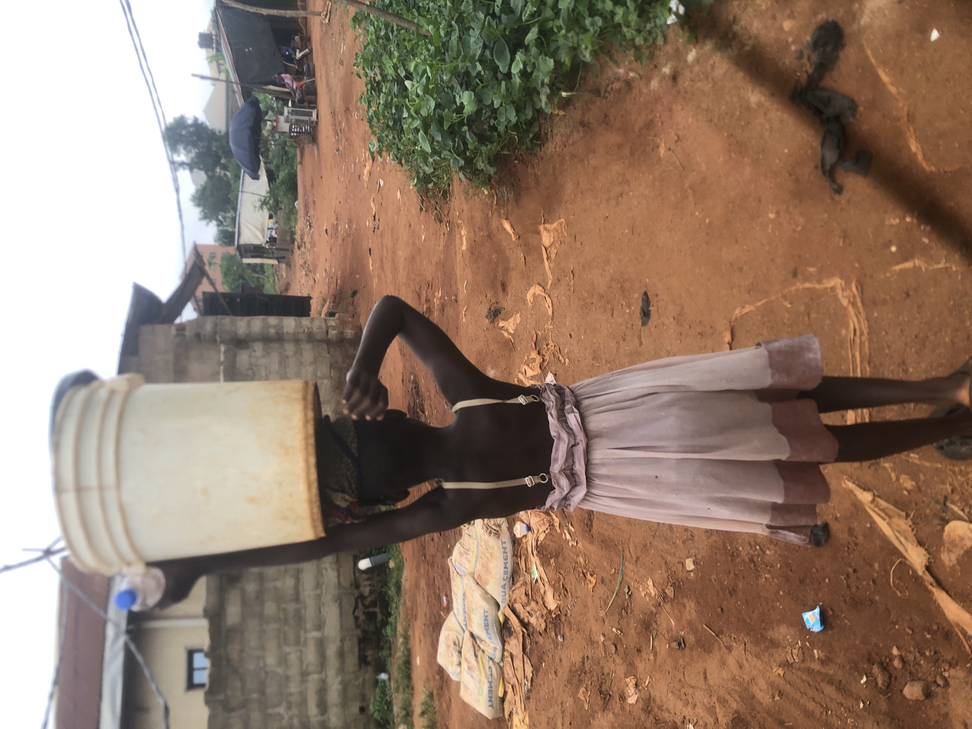 An outdoor, eye-level shot in Ukwu-Abwa, Nigeria, shows a young girl carrying a large, off-white bucket on her head, positioned slightly right of the frame and facing left. She wears a dark top with thin straps, a light-colored ruffled skirt or dress, and a dark head covering. A small plastic bottle with a blue cap is attached to the side of the bucket.
The setting is an unpaved area with reddish-brown dirt, indicating a rural or semi-urban environment. The ground is scattered with debris, including plastic bags and wrappers. Several bags labeled "BUA CEMENT" are visible on the ground behind the girl. To the left, a lush green bush adds a touch of natural vegetation.
In the background, various simple structures and buildings are visible, some made of grey concrete blocks and others appearing more temporary with dark tarpaulins or sheets. A dark blue open umbrella is notable in the mid-ground. Overhead, power lines span across a light, overcast sky, suggesting it is daytime with diffused lighting and no harsh shadows. The scene depicts a common daily activity of water transportation in a developing community.