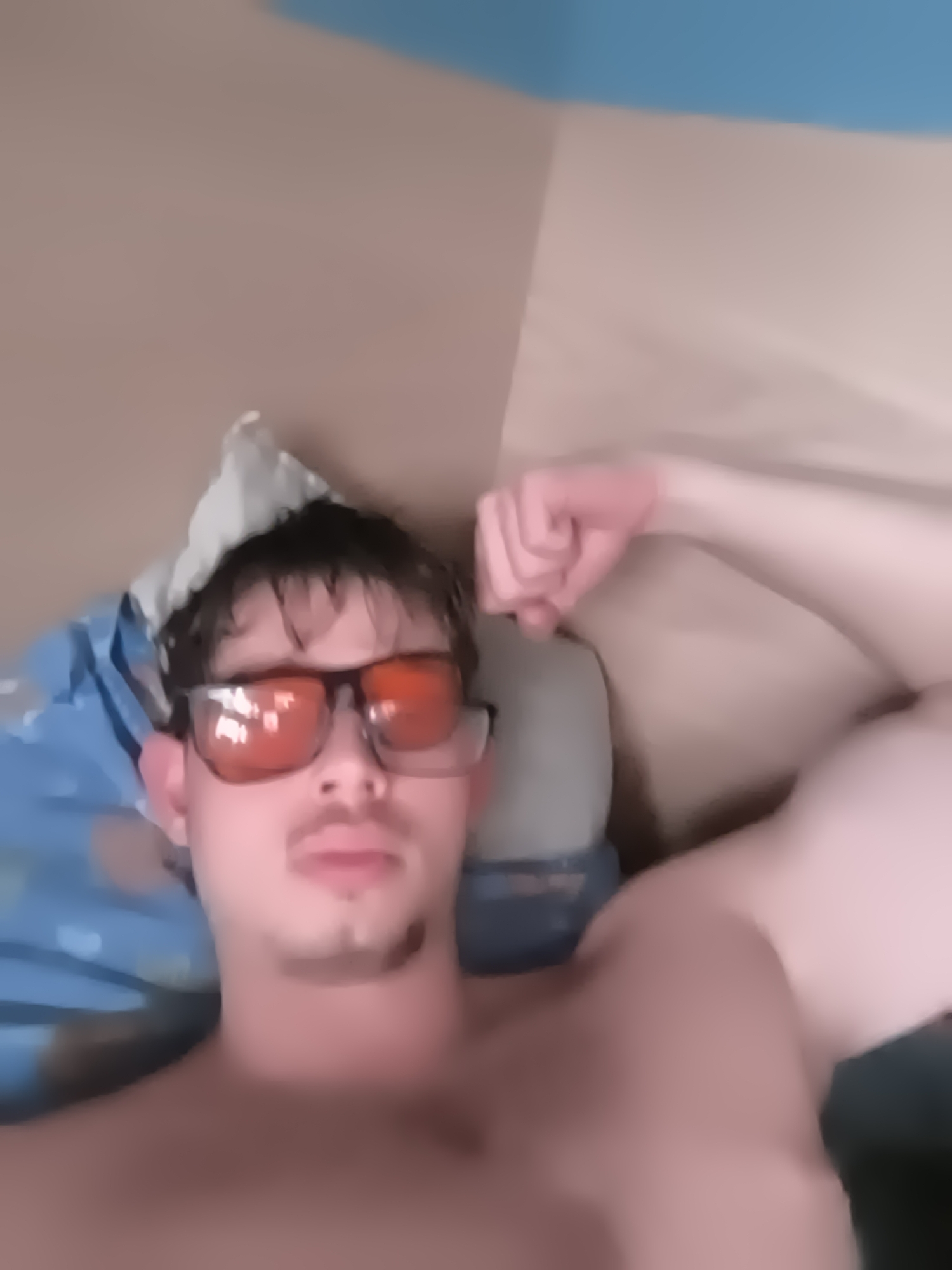 A blurry, top-down, close-up shot features a young man lying down indoors, possibly on a bed or couch. He is captured from the chest up, with his right arm bent and flexed, showcasing his bicep. The man has dark, shaggy hair, a mustache, and a goatee. He is wearing dark-framed sunglasses with orange-tinted lenses that obscure his eyes.
The background shows a plain, light brown or beige wall, with a strip of light blue at the top, indicating an upper wall or ceiling area. To the left of the man, a blue pillow or blanket with a faint pattern is visible. A lighter, possibly white, pillow or crumpled fabric is positioned behind his head. The lighting is soft and ambient, suggesting an indoor setting without strong natural light. The overall image is significantly out of focus, giving it a soft, indistinct quality. No text is visible in the image.