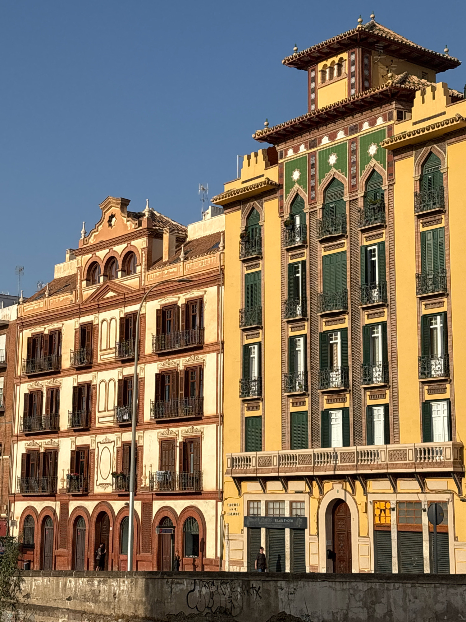 The image depicts the facades of two adjacent buildings in Málaga, Spain, along the Guadalmedina River. The building on the right is a vibrant yellow with intricate green and red tiled decorative elements and numerous arched windows with green shutters. It features ornate balconies with wrought-iron railings. The building on the left is a lighter shade of cream with terracotta accents, also featuring multiple arched windows with dark shutters and balconies. The lower levels of both buildings reveal shopfronts and doorways. People are visible on the sidewalks in front of the buildings, and a low concrete wall with graffiti runs along the foreground, separating the buildings from the riverbed. The sky is a clear, bright blue.