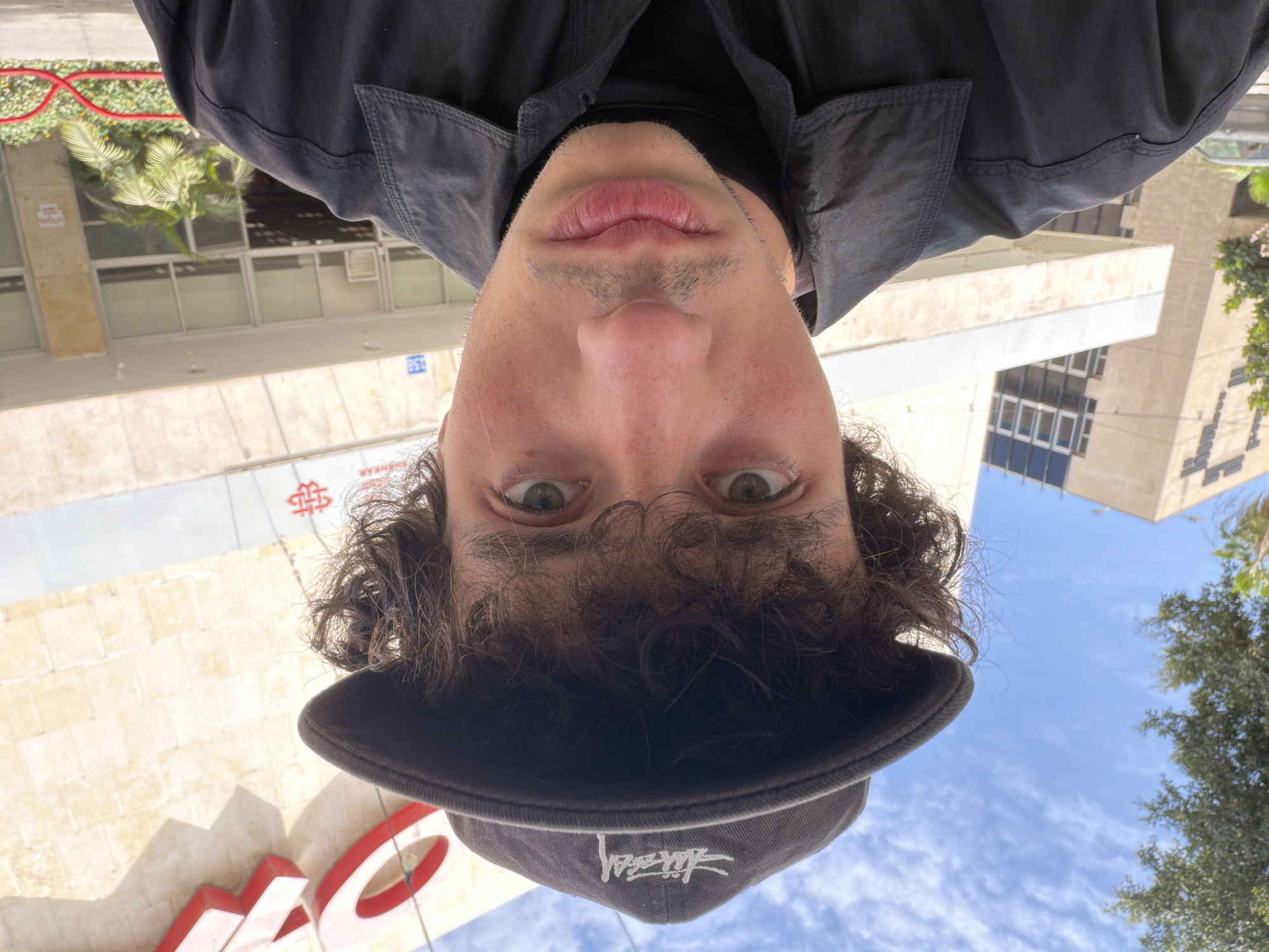 A man wearing a black jacket and a black Stussy hat is looking at the camera. He is outside in Tel Aviv, Israel, based on the Hebrew writing on the buildings. The sky is visible above him.
