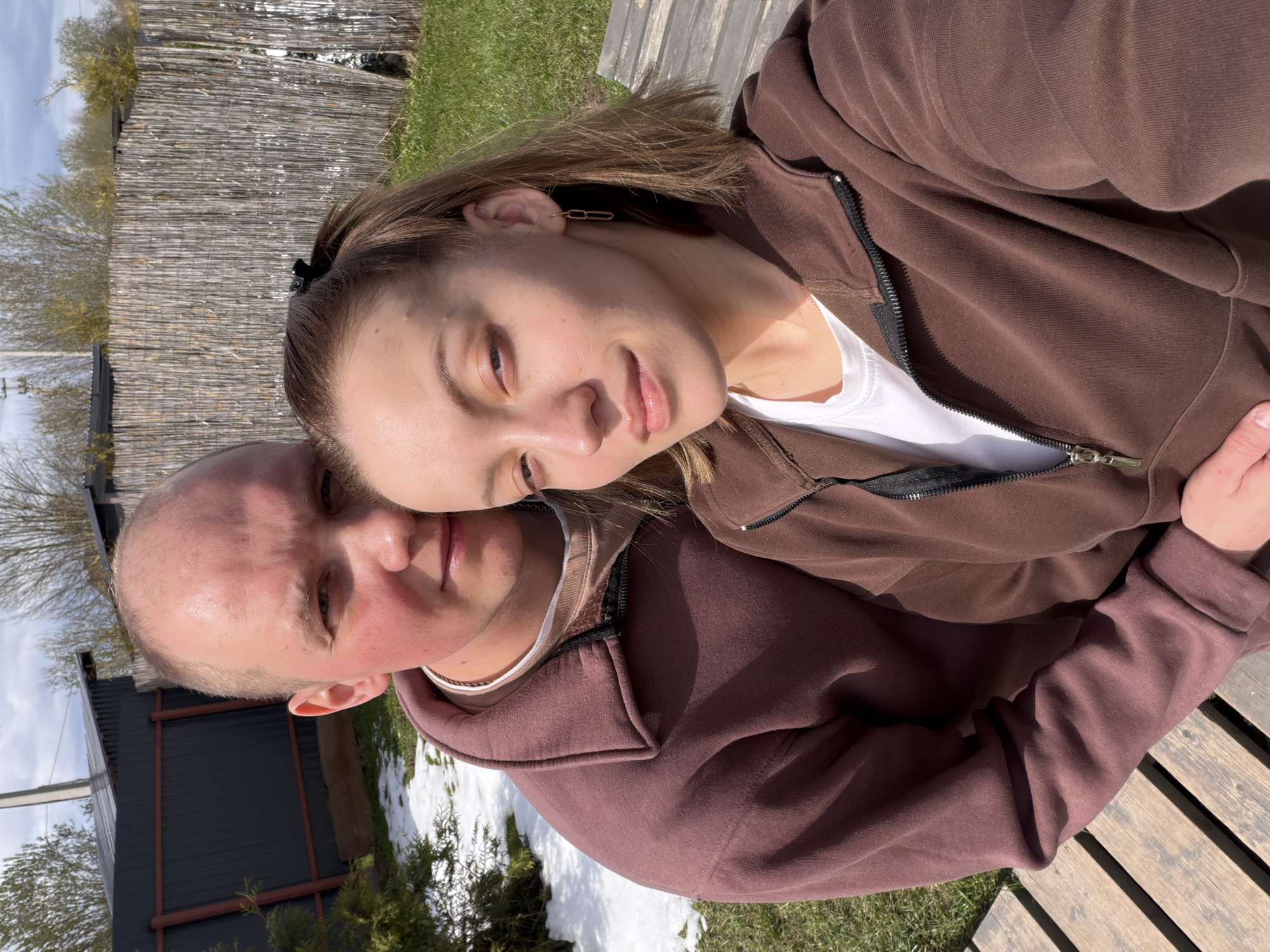 A horizontal outdoor photo captures a couple on a bright, sunny day, likely in early spring, indicated by the green grass interspersed with patches of melting snow. The woman, with medium-length brown hair secured by a black claw clip, leans her head gently against the man's shoulder. She wears a brown zip-up hoodie over a white t-shirt and sports a golden chain-link earring in her right ear, looking towards the camera with a soft smile. The man, with short, light brown hair, wears a slightly darker, reddish-brown zip-up hoodie and looks towards the camera with a neutral expression. They appear to be sitting on a wooden slatted bench, which is partially visible in the lower right.
The background features a rustic woven wooden fence or wall. Behind the man, a dark-colored, simple building is visible. Sparse trees with emerging green leaves stand against a clear blue sky dotted with white clouds. The scene suggests a relaxed, casual setting in a rural or semi-rural area, consistent with the provided location context of Бунятино, Russia. There is no visible text in the image.