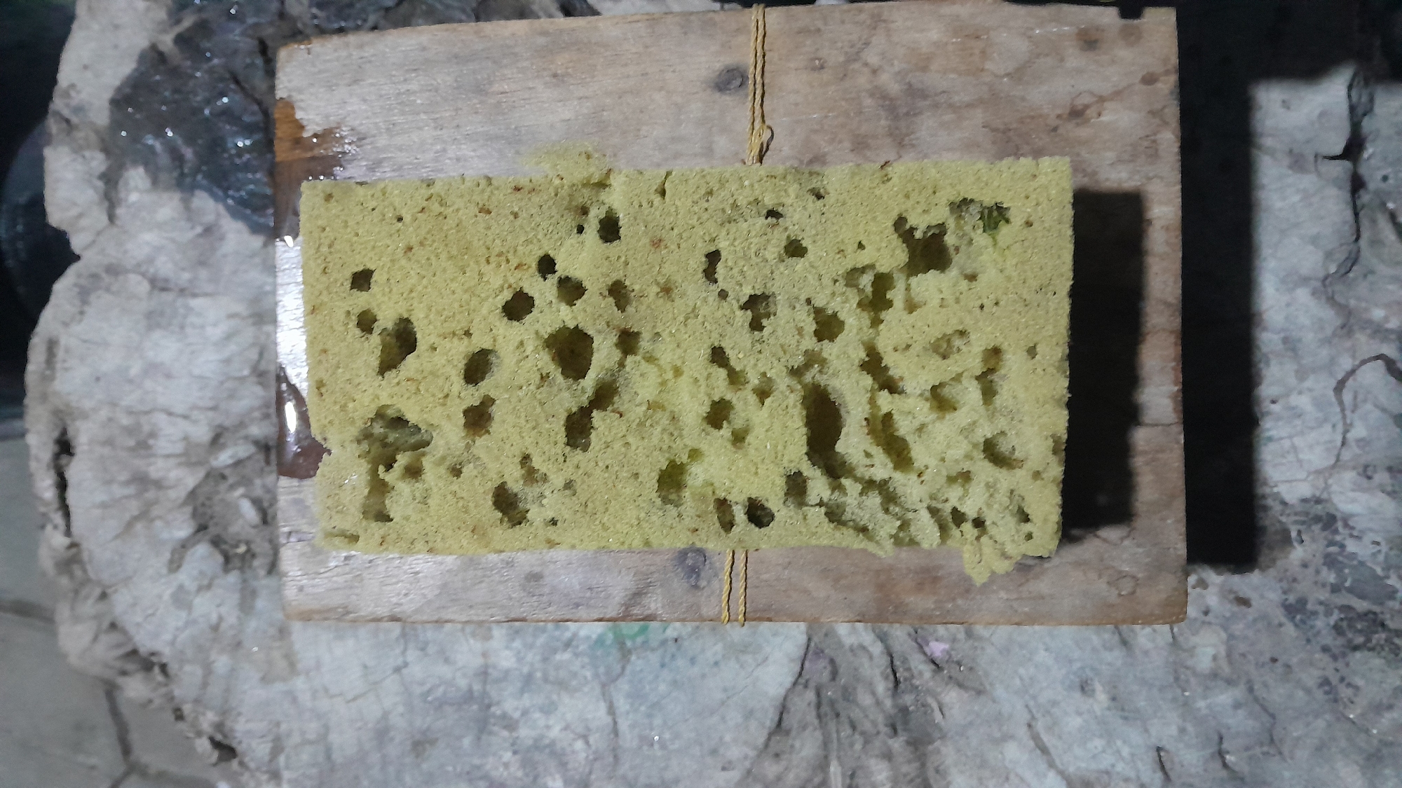A close-up, overhead shot shows a yellow sponge resting on a wooden plank. The sponge is rectangular with numerous irregular holes throughout its surface. The wood plank is stained and has two pieces of yellow twine wrapped around it, securing the sponge. The background consists of a grey, textured surface, possibly concrete or stone, and a darker, wet-looking area with reflections. The overall scene appears to be outdoors or in a dimly lit space. There are no people or animals visible in the image. The image provides no specific information about the location in the city of Sumberarum, Indonesia.