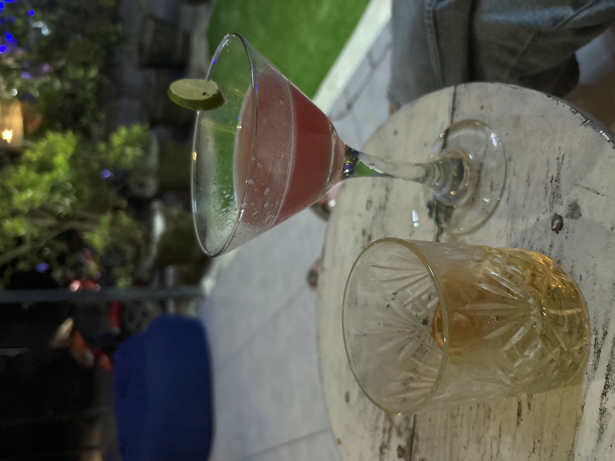 On a weathered, round, white table, sits a pink cocktail in a martini glass garnished with a lime wedge. Beside it, a cut-crystal glass holds a golden liquid, possibly whiskey. The setting is outdoors, with a glimpse of greenery and patio tiling in the background. A person's leg is visible near the table. The city of Kuta Selatan, Indonesia, is not explicitly depicted, but the outdoor setting and potential tourist presence suggest a location consistent with the area.