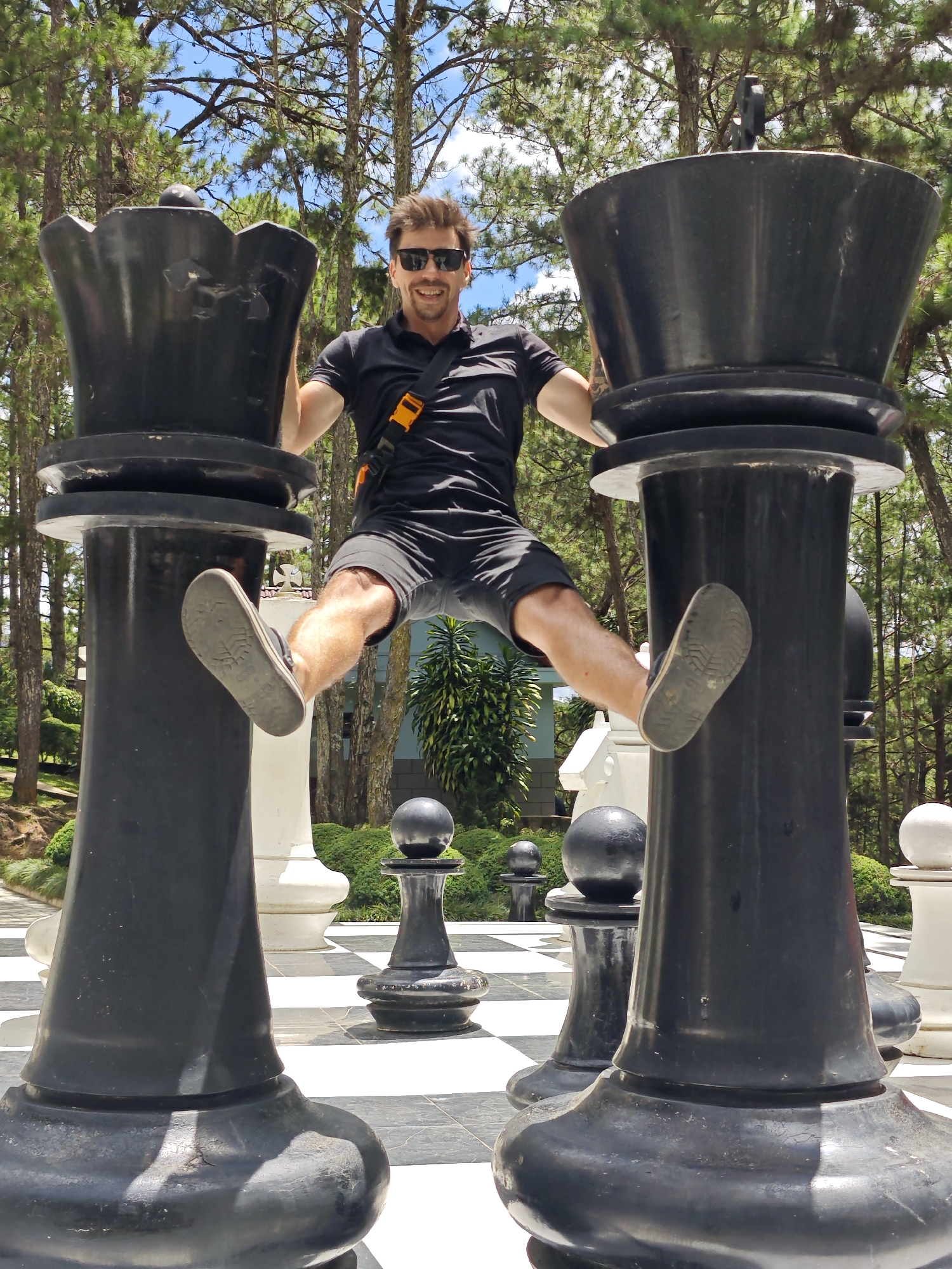 A man is captured mid-air, playfully straddling two giant black chess queen pieces on a checkered outdoor board. He is wearing a black polo shirt, black shorts, sunglasses, and a strap across his chest with an orange buckle. His legs are spread wide, with one foot resting on each chess piece. The setting appears to be a park or garden, with tall pine trees and lush greenery in the background. The sky is blue with scattered clouds, suggesting a pleasant daytime with sunny weather. The giant chess pieces indicate the location is likely a tourist attraction, possibly a park or resort. The man's pose and expression suggest a lighthearted and adventurous mood. The overall scene is bright and vibrant, conveying a sense of fun and relaxation. The location is Lam Vien - Da Lat, Vietnam.