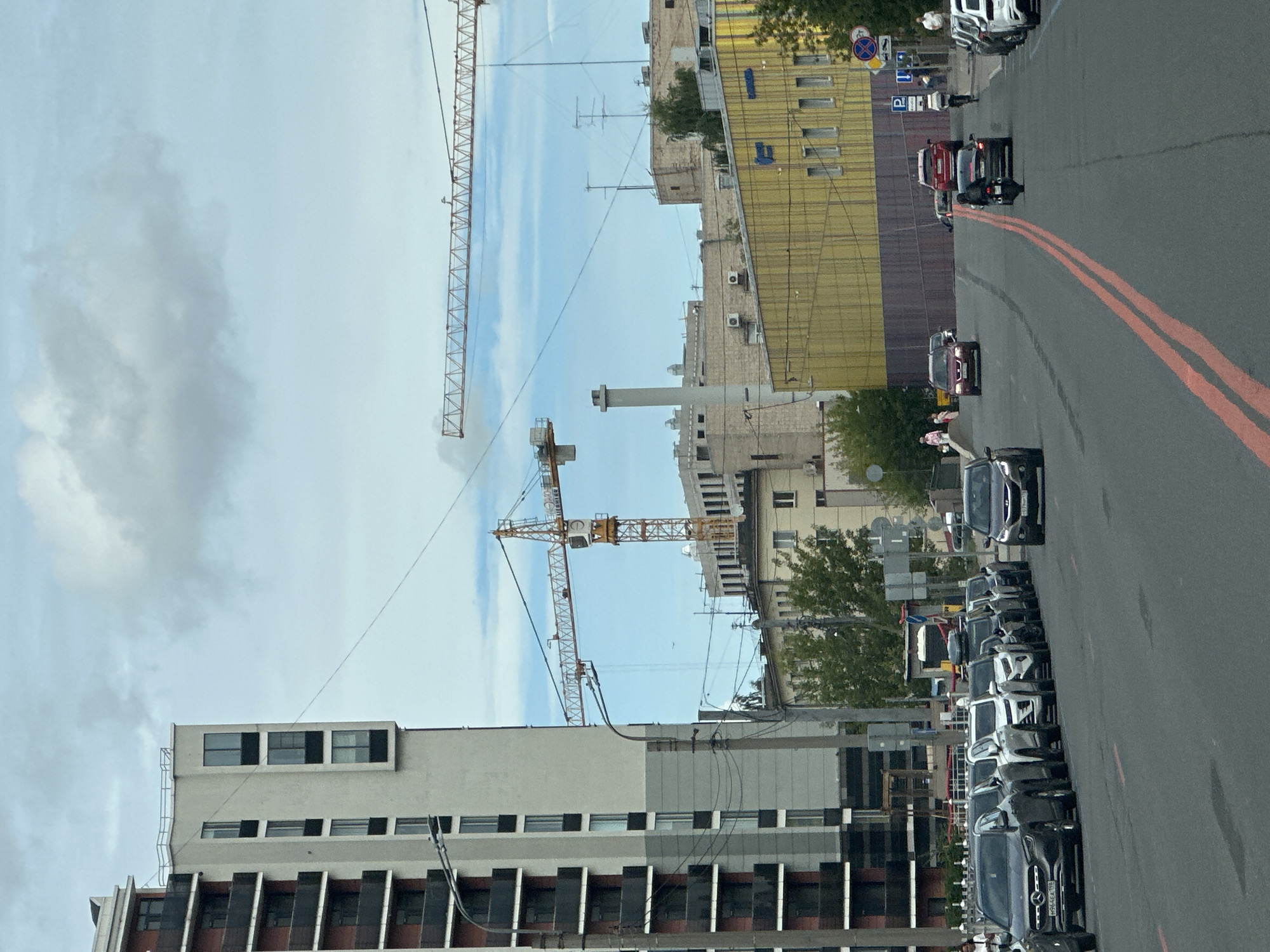 The image captures a street scene in Saint Petersburg, Russia, under a cloudy sky. A multi-story residential building with dark balconies is on the left. Further down the street, construction cranes are visible, indicating ongoing building projects. Several cars are parked along the roadside and driving on the asphalt road, which features double red lane markings. A motorcycle is also seen on the road. In the background, various buildings, including a yellow-colored structure, are present. People are walking on the sidewalk and near the buildings. The overall environment suggests an urban setting with a mix of residential, commercial, and construction areas.